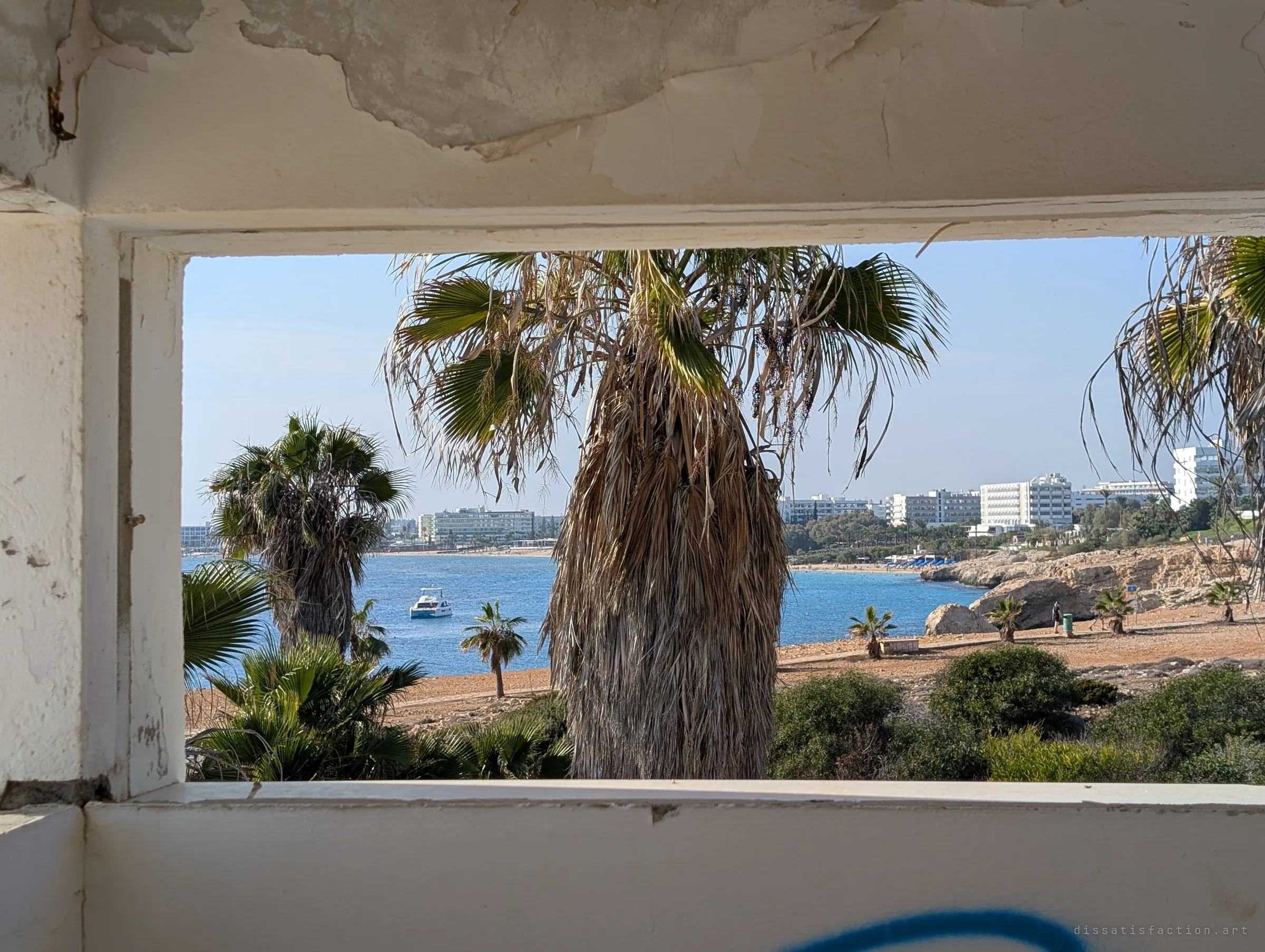 A peeling concrete window opening frames palm trees, a turquoise bay and a line of seaside hotels in the distance. The ruin looks out on the busy coast it was built to serve.