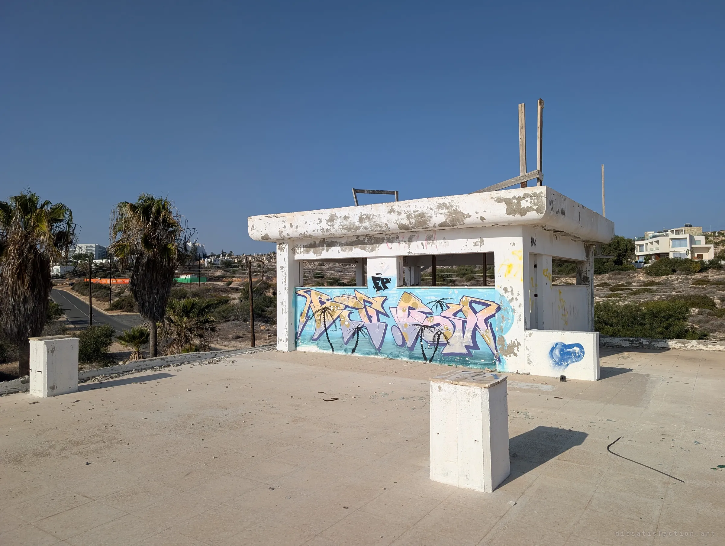 A small open rooftop kiosk, its wall covered in a pastel graffiti piece with painted palm trees, stands on an empty tiled roof under a deep blue sky. Broken timber posts stick up from its flaking concrete canopy.