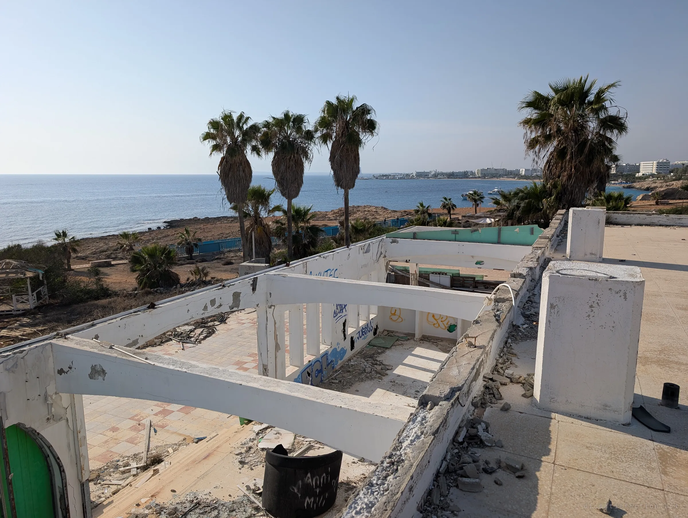 From the roof, the gutted upper floor opens below, with bare beams, graffiti and rubble on checkered tiles, while tall palms and the open sea fill the horizon. The building sits right on the shoreline, stripped to its frame.