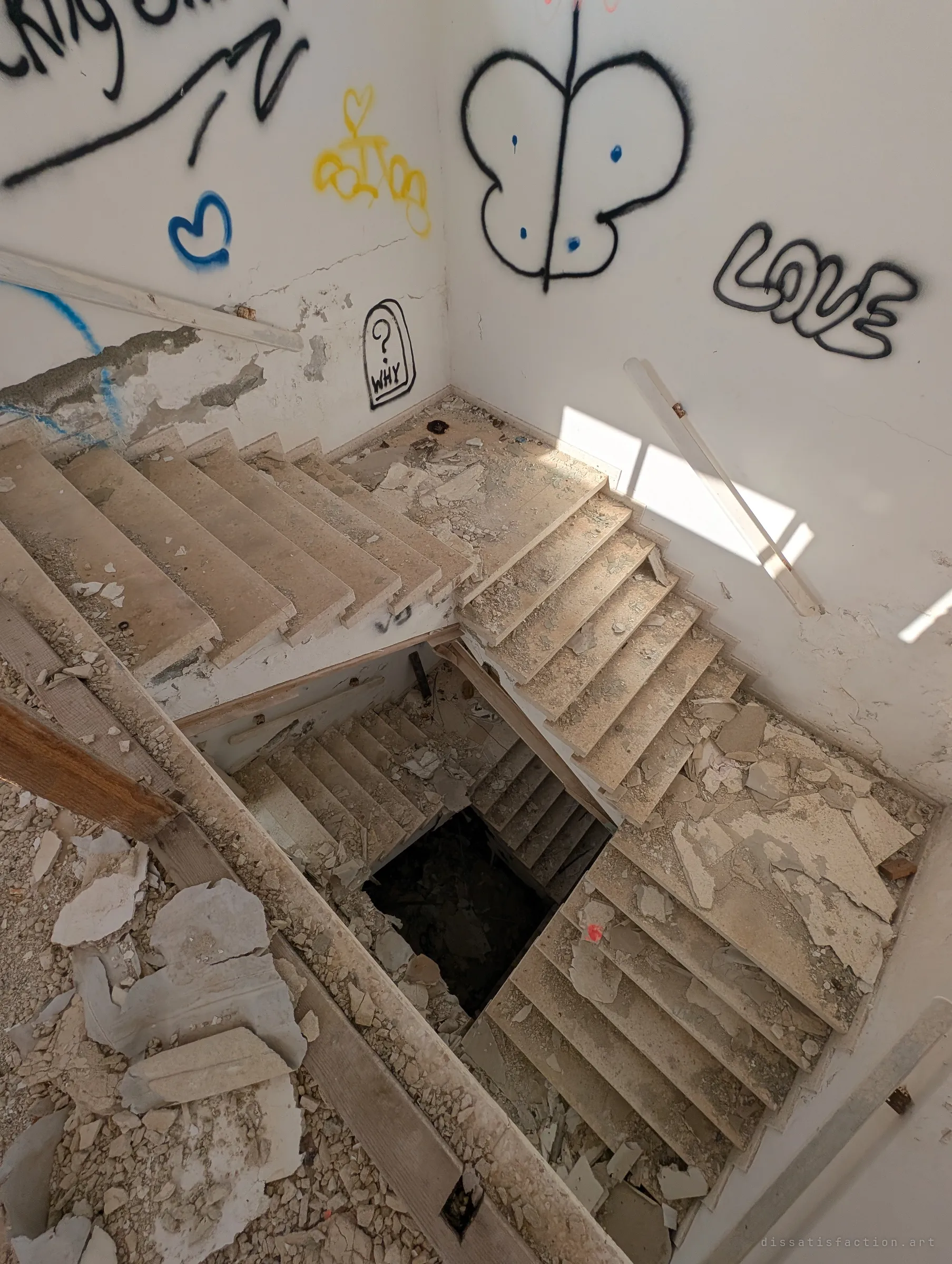 Looking straight down a debris-strewn stairwell, flights of broken stone steps spiral into a dark void. Graffiti hearts, a butterfly and the word love cover the sunlit walls above.