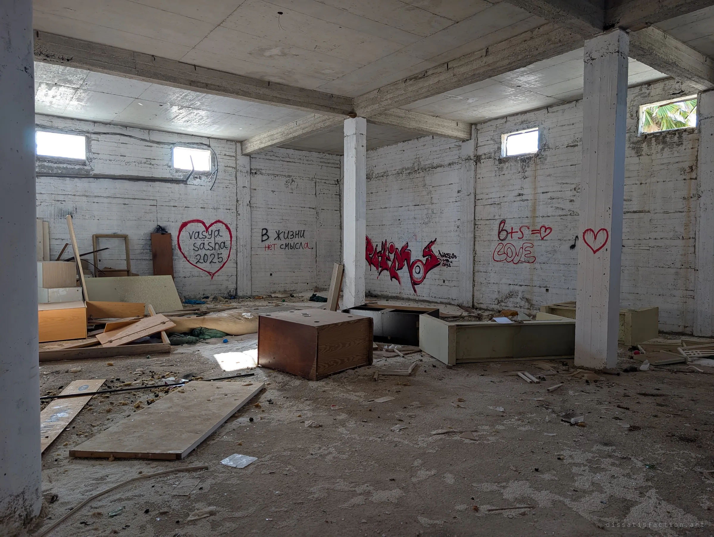A basement-like room with small high windows holds tipped-over cabinets and scattered panels. Red love-heart graffiti and a line of Russian text about life having no meaning mark the pale walls.