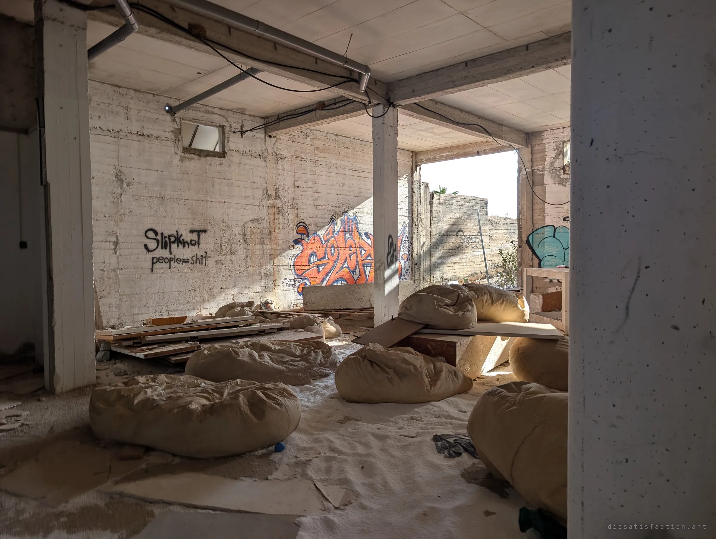 Sunlight cuts through an open side of a concrete room full of slumped beanbags half buried in foam beads. An orange graffiti piece and a band name scrawled in black cover the back wall.