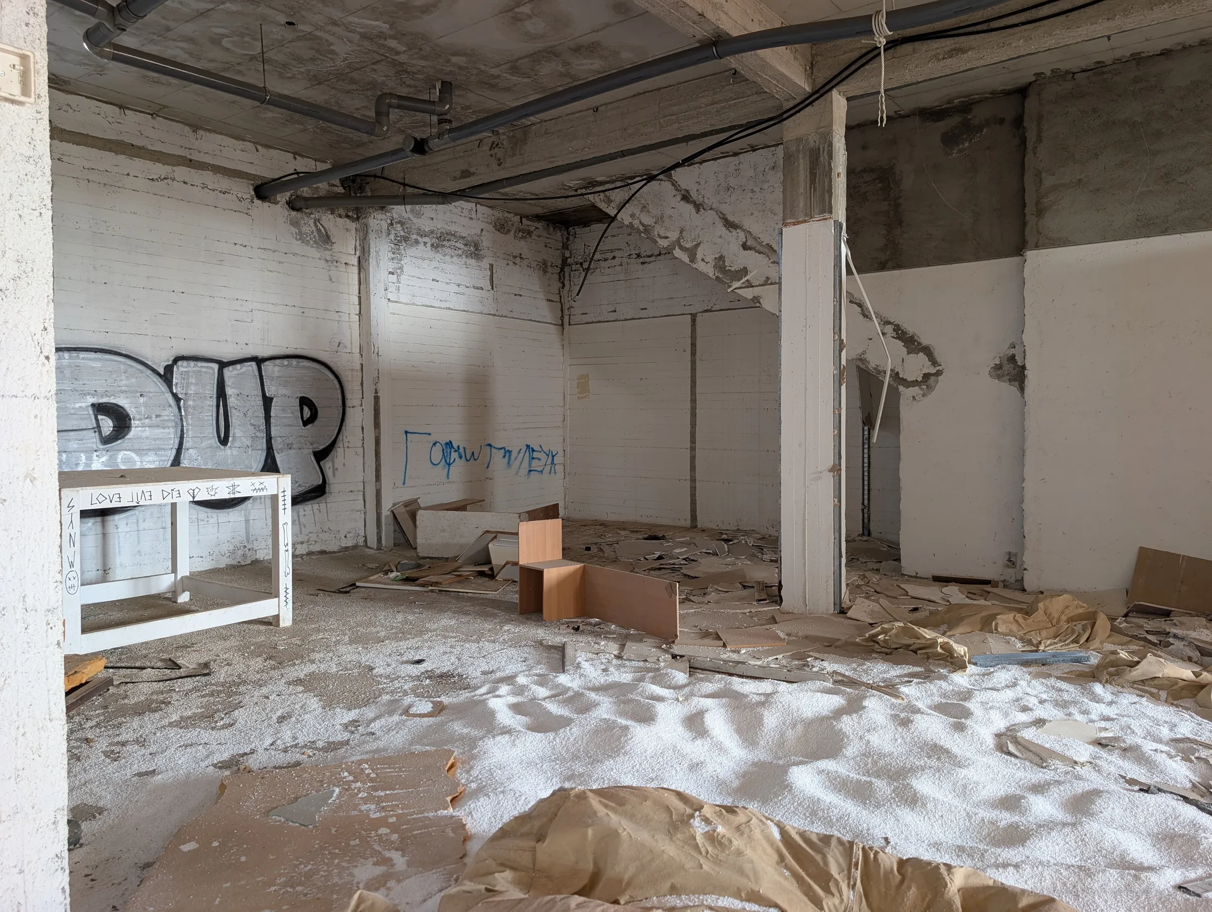 A stripped interior under exposed pipes, with a white table marked in runes, broken furniture and a floor drifted with white foam beads like snow. A large silver graffiti tag fills the far wall.