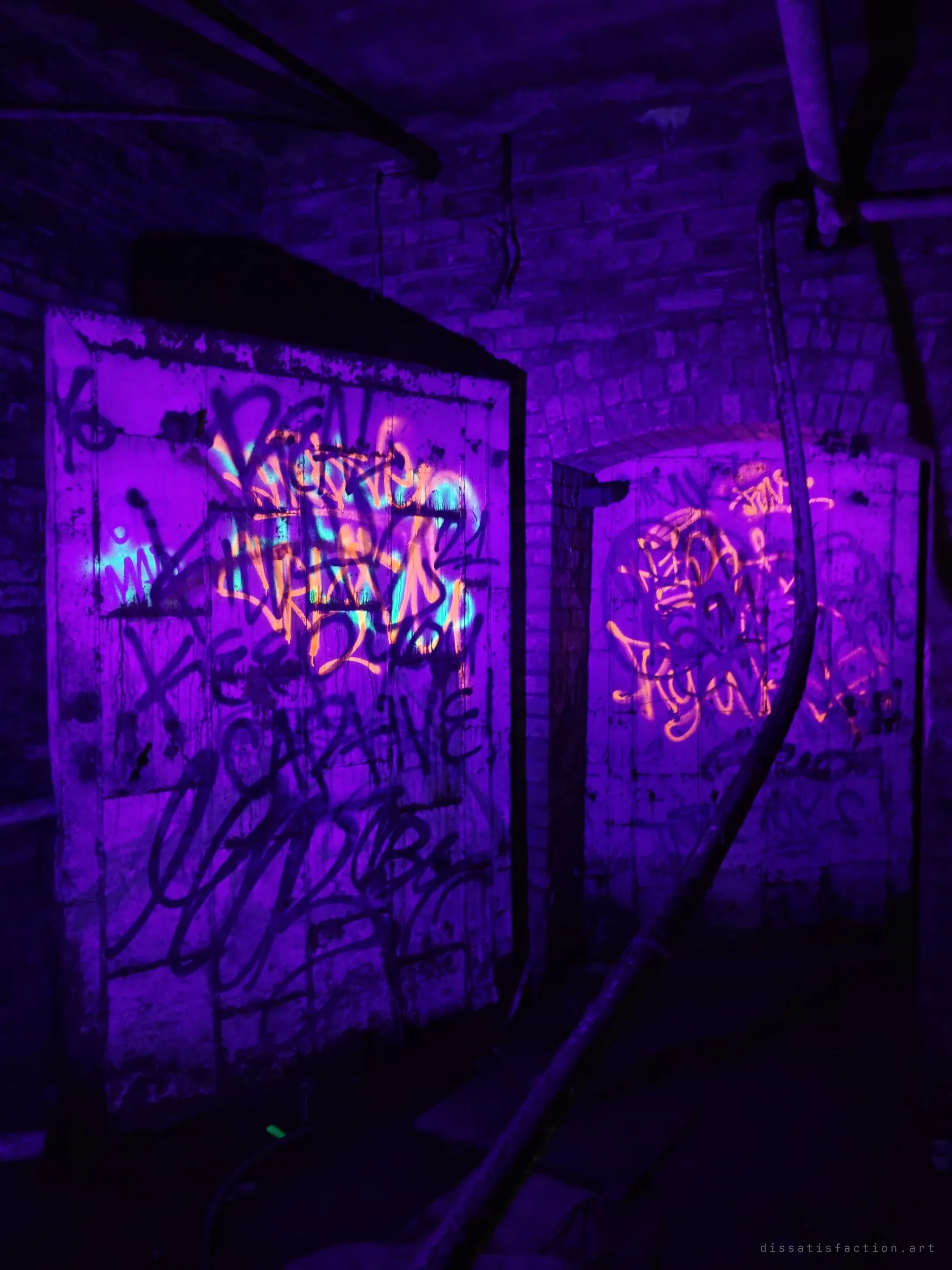Two graffiti-covered doors in a brick basement glow under ultraviolet light, the fluorescent tags burning orange and cyan against deep violet.
