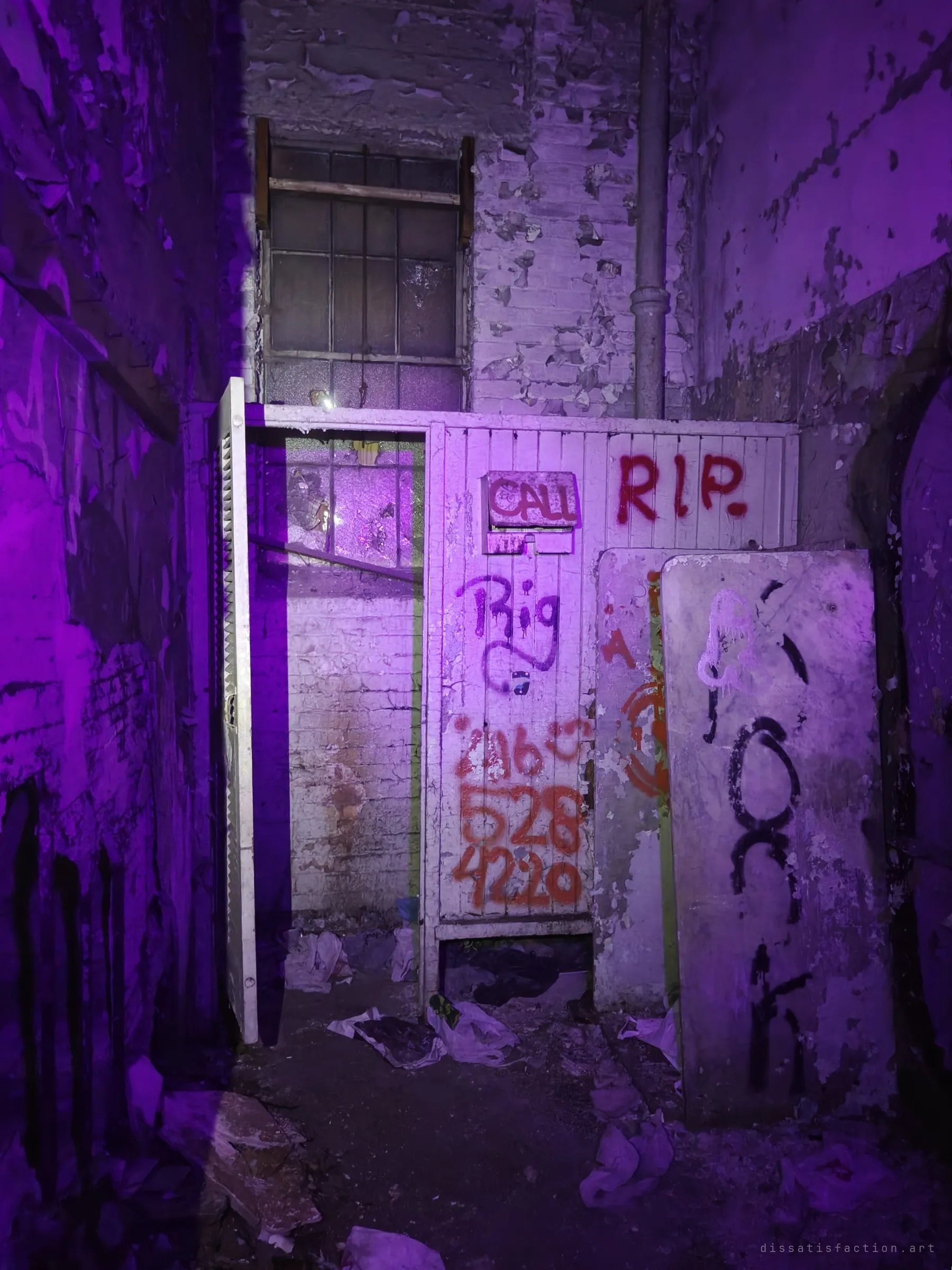 A cramped, violet-lit corner with peeling white paint, a wooden partition covered in spray-painted messages and an old mattress leaning against the wall beneath a barred window.