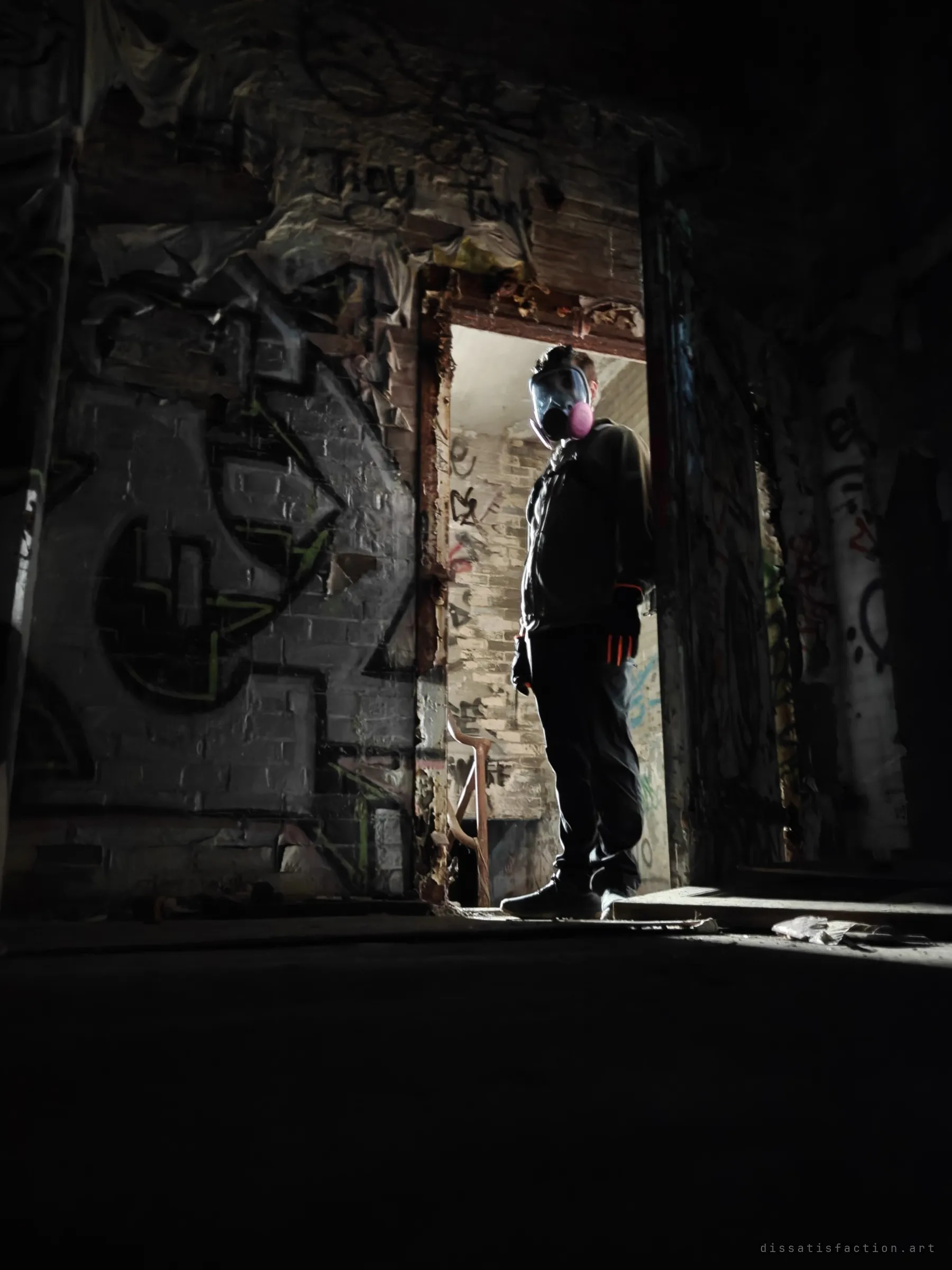 A figure in a full-face respirator stands silhouetted in a bright doorway, framed by dark graffiti-covered brick and a stairwell beyond.