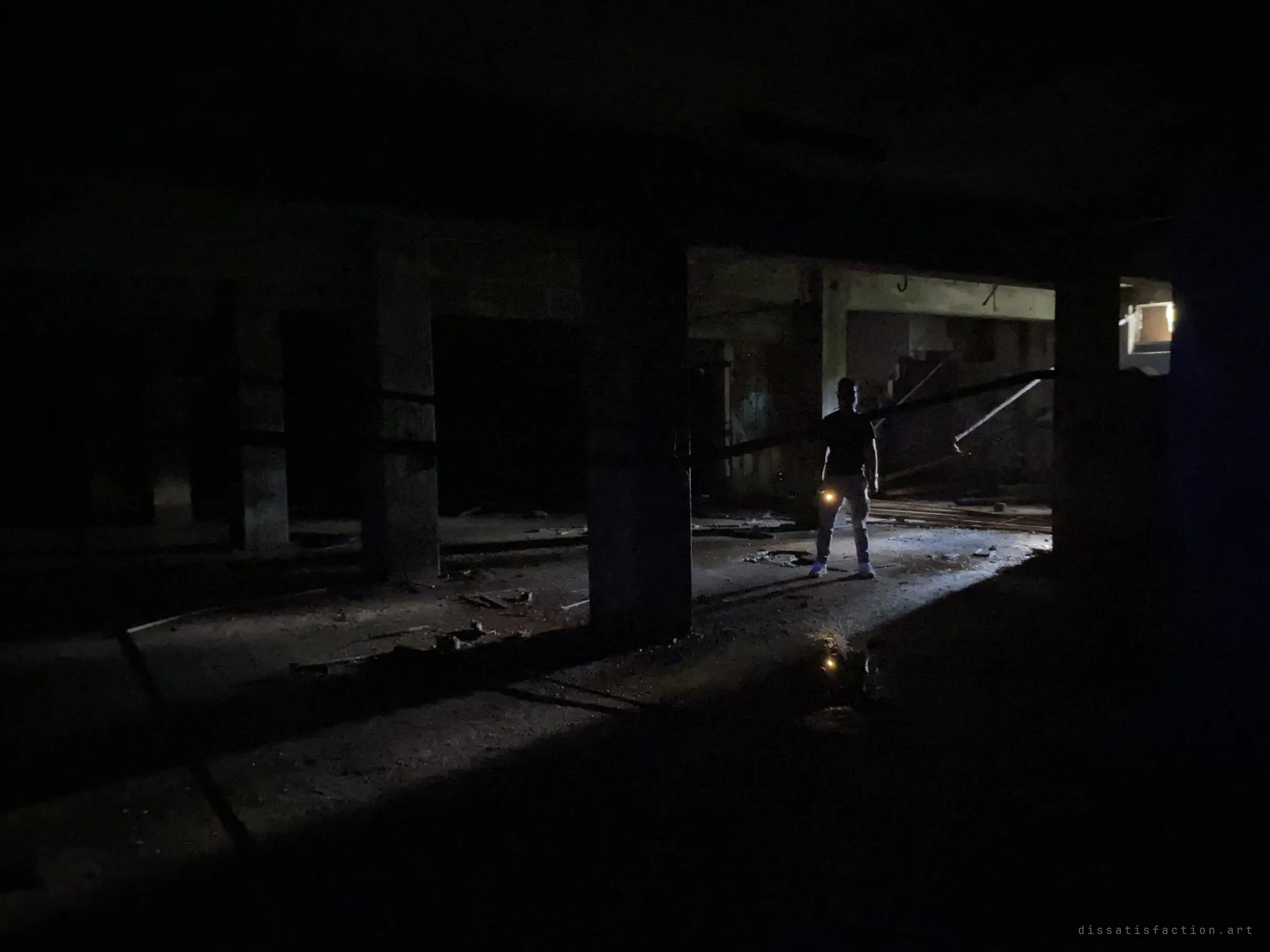 A lone figure holding a small light stands in a pool of daylight among the heavy concrete columns of a dark, debris-strewn ground floor.