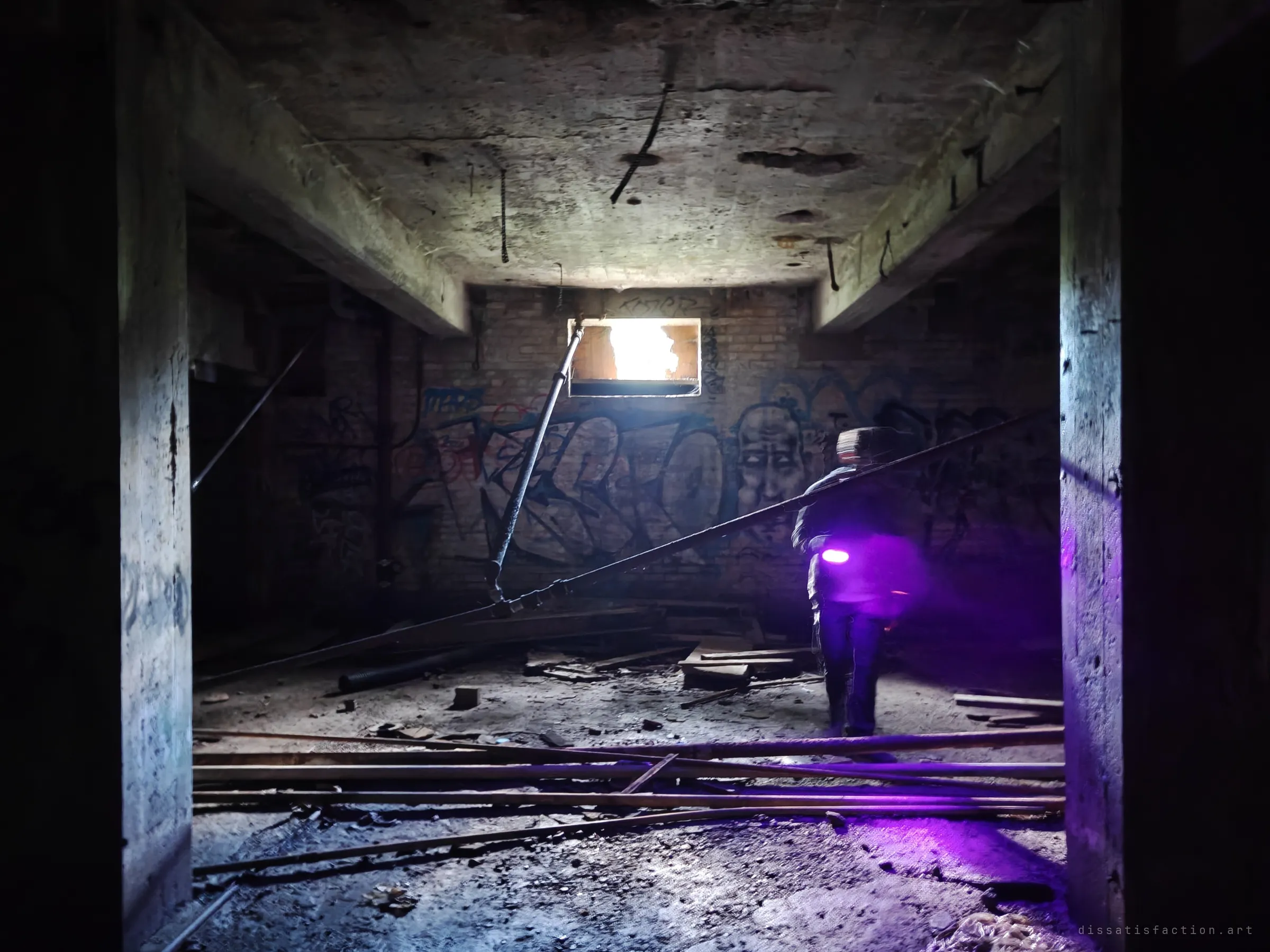 A low concrete room with exposed rebar, a single bright window and fallen pipes across the floor, where an explorer with a violet light walks past a painted face in the graffiti.