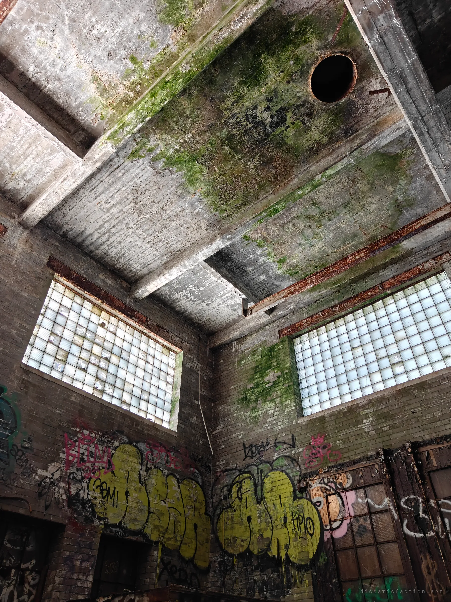 Looking up into a tall mill room: moss-streaked concrete ceiling with a round chute hole, rusted steel beams and two walls of glass-block windows above bright graffiti.