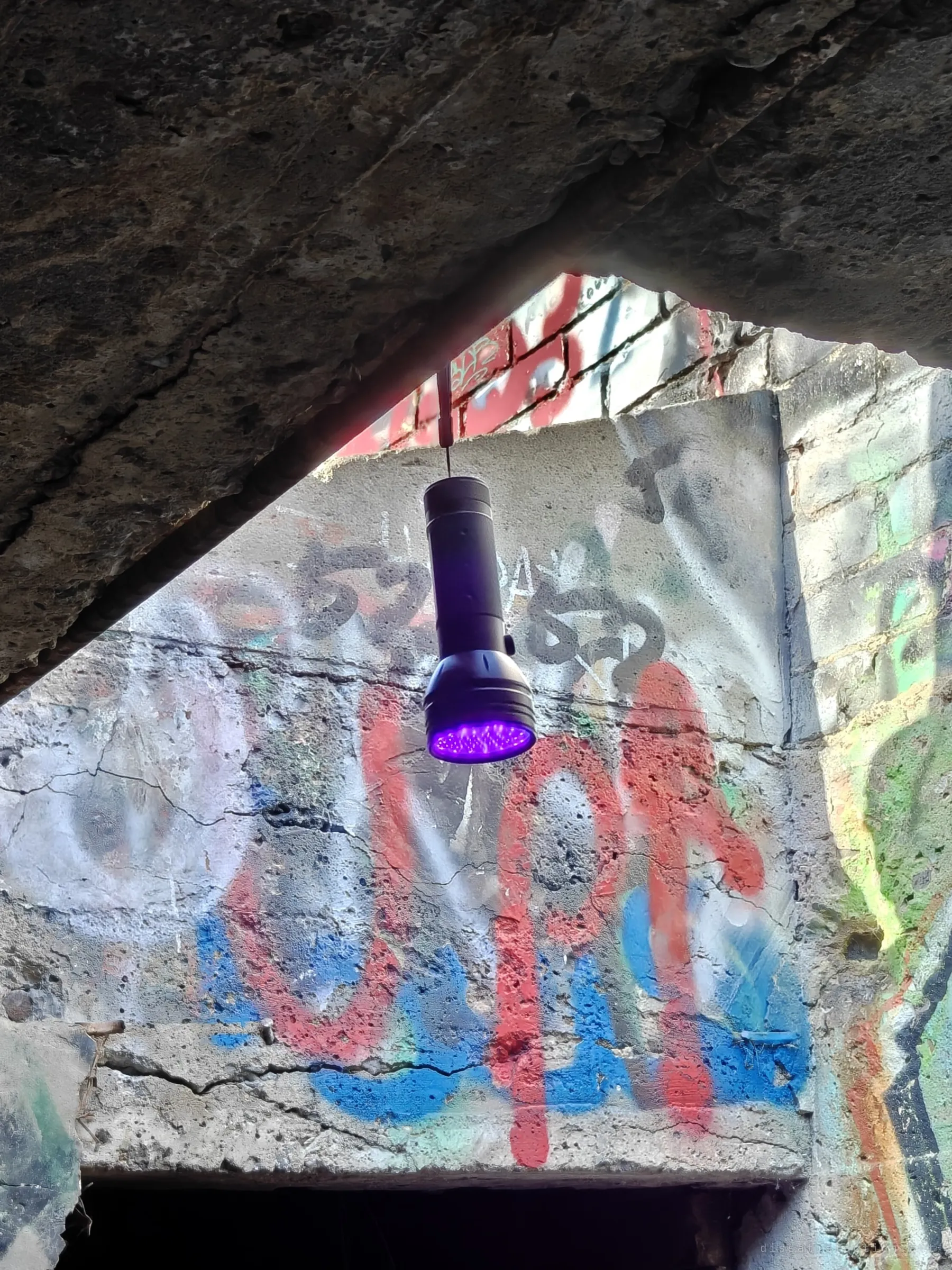A UV flashlight hangs from a strap beneath a broken concrete slab, its violet LEDs glowing against a wall of faded graffiti over a dark doorway. A small setup detail from the night side of the visit.