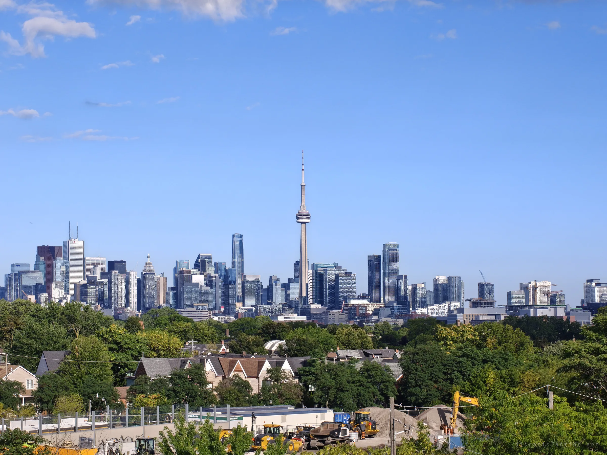 A wide view of a downtown skyline and its tall observation tower under a clear sky, seen over tree-lined houses and a construction yard. The view from the roof is part of why the place draws people back.