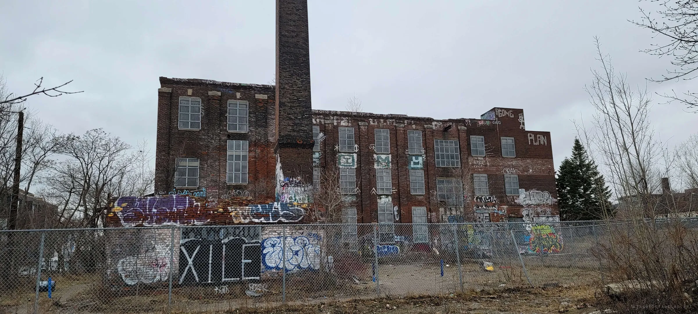 A graffiti-covered brick factory with a tall square chimney stands behind a chain-link fence under a grey sky. It sets the scene: a large, long-abandoned industrial complex.