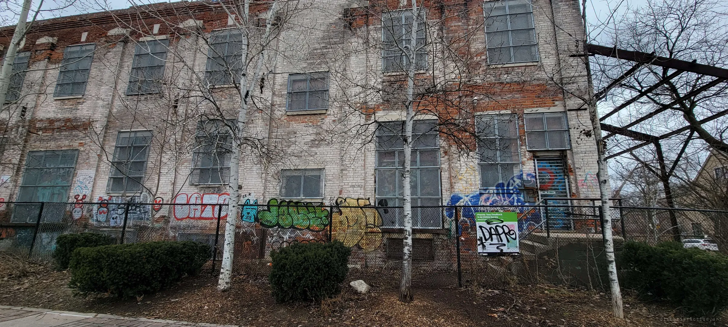The pale, peeling brick side of the factory seen from the sidewalk through young birch trees and a black fence tagged with graffiti. A defaced public notice hangs on the fence.
