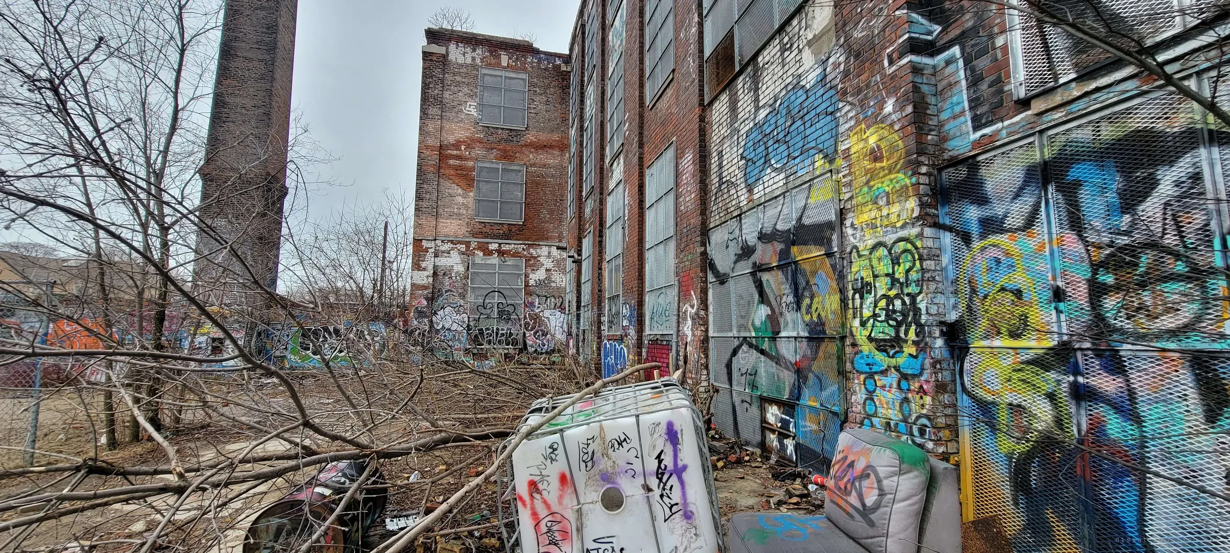 A littered yard runs along a wall of graffiti-covered steel mesh toward a narrow brick wing, with the chimney rising at left. A tagged plastic tank and a discarded armchair sit among fallen branches.