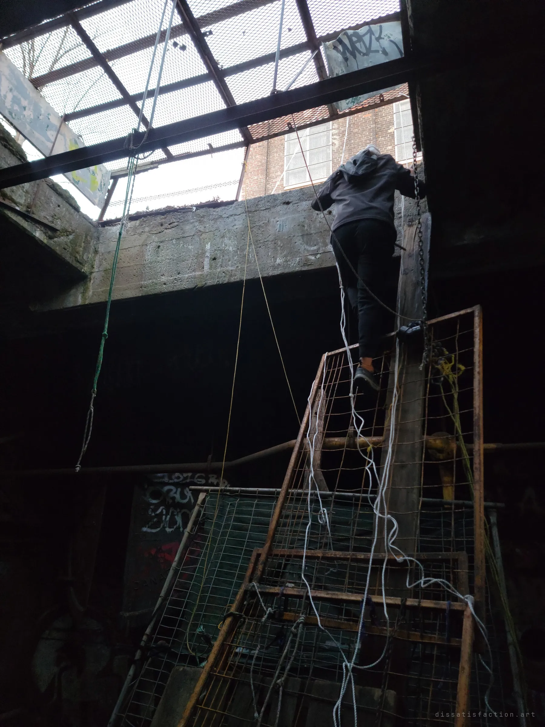 Seen from below, an explorer climbs a makeshift ladder of rusted mesh and rope up through an opening under a steel grate. It shows the improvised way in and out through the roof.