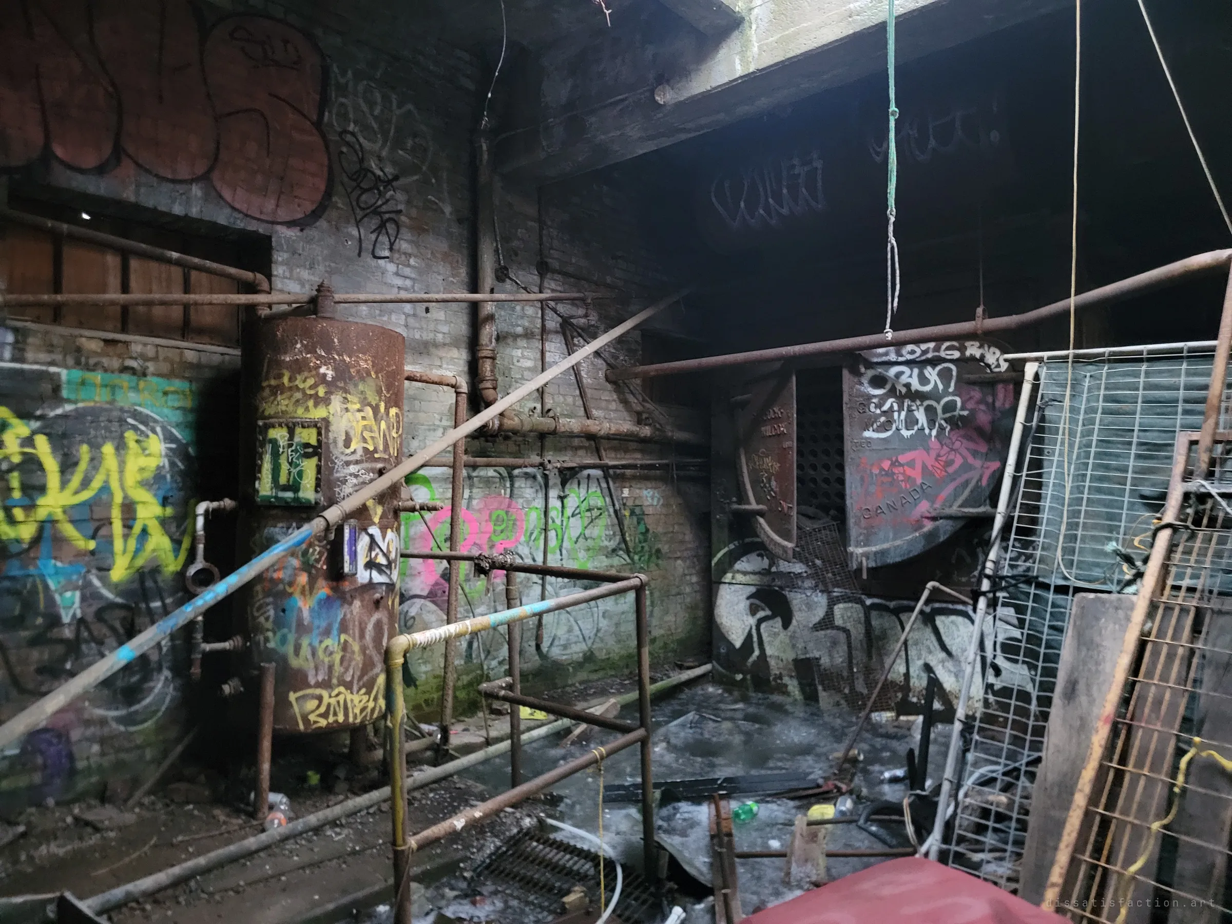 A dim boiler room with a rust-streaked tank, tangled pipes and an old furnace door, every surface covered in graffiti. Ropes hang from the ceiling above scattered debris and frozen puddles.
