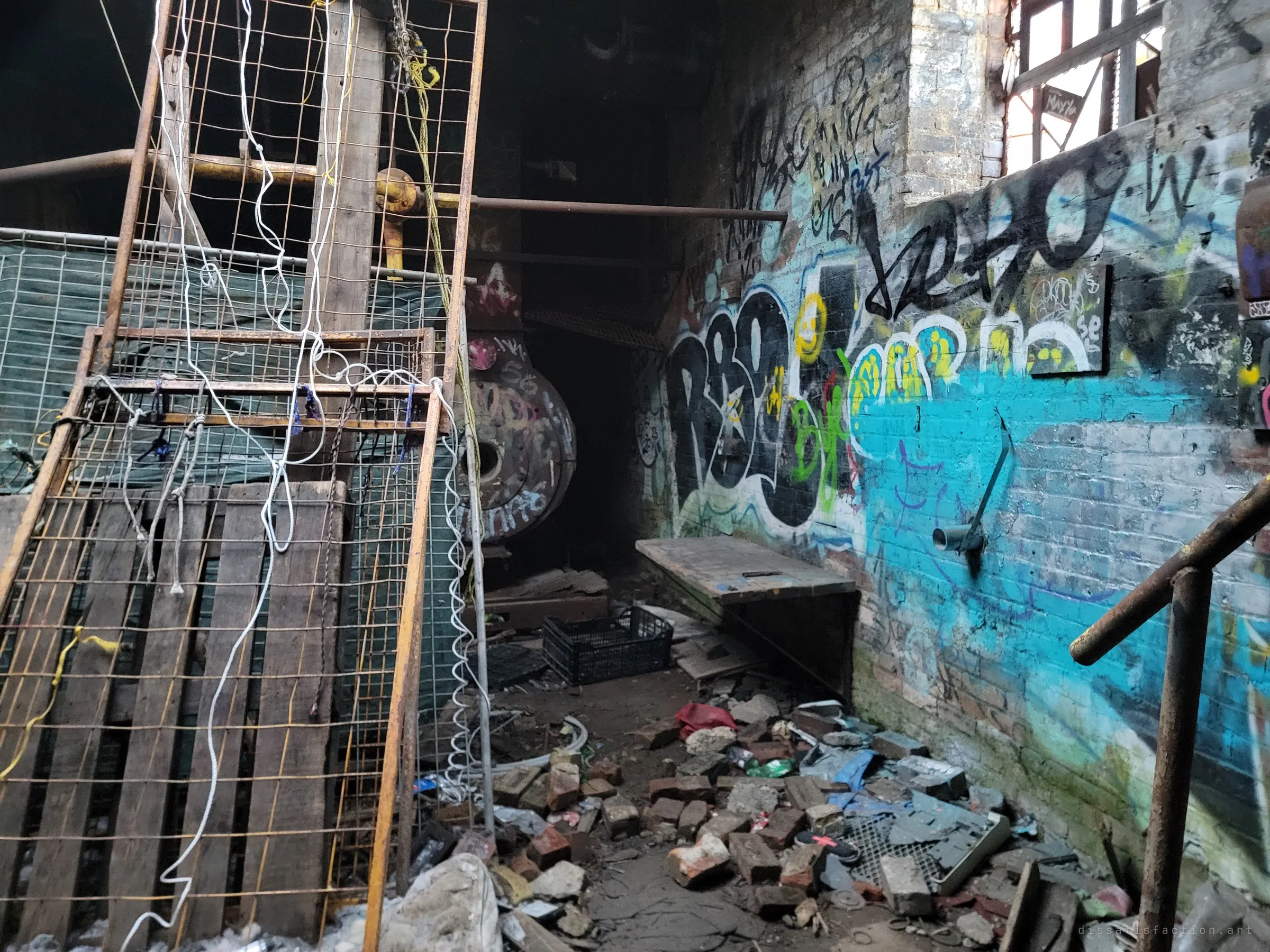 Rope-tied mesh panels and pallets lean beside a bright turquoise graffiti wall, with rubble and broken bricks on the floor. A large round boiler hides in the dark behind them.