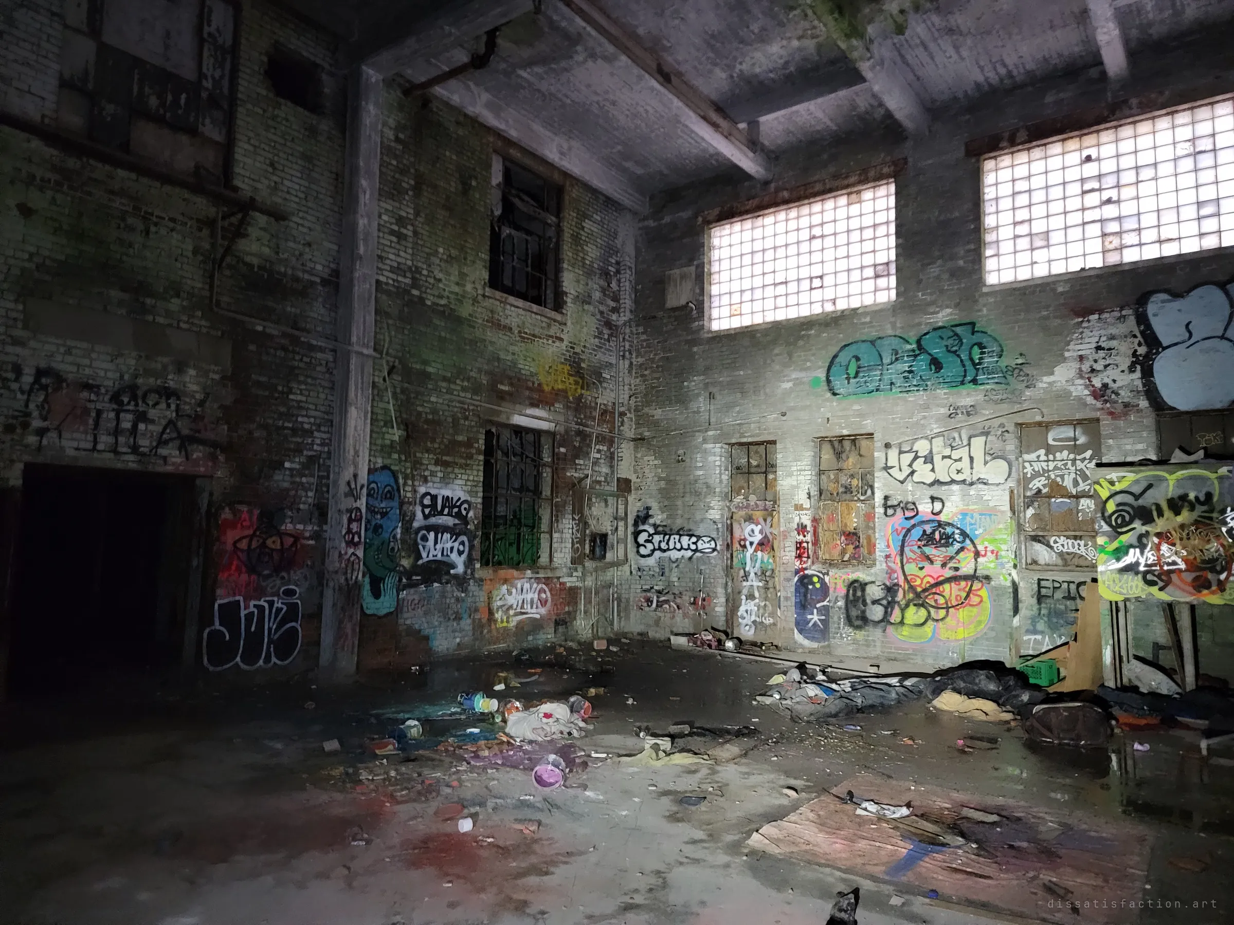 A tall, empty brick hall lit by high glass-block windows, its walls layered with tags and pieces. Paint cans, bedding and debris are strewn across the wet concrete floor.