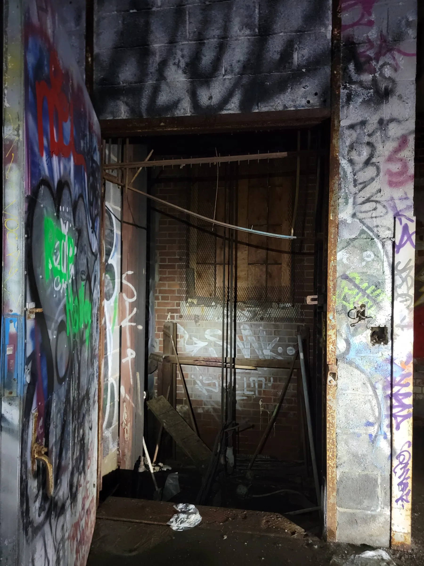 A heavy graffiti-covered door opens onto an old freight elevator shaft, lit by a flashlight. Broken boards and cables hang inside against the brick back wall.