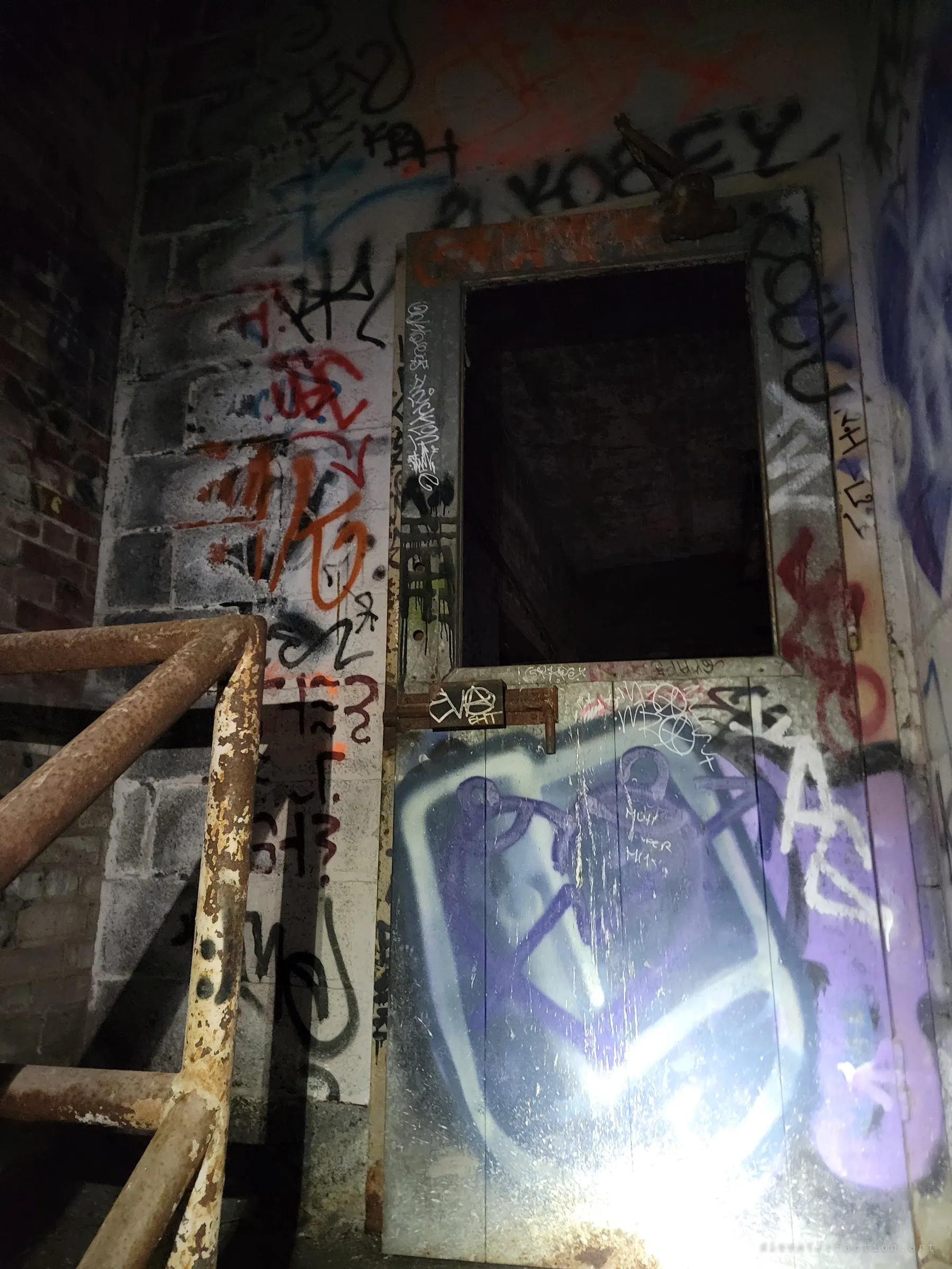 A rusted stair railing climbs past a steel door buried under layers of graffiti tags, its upper panel open onto darkness. The flashlight glare shows how thoroughly the stairwell has been claimed by visitors.