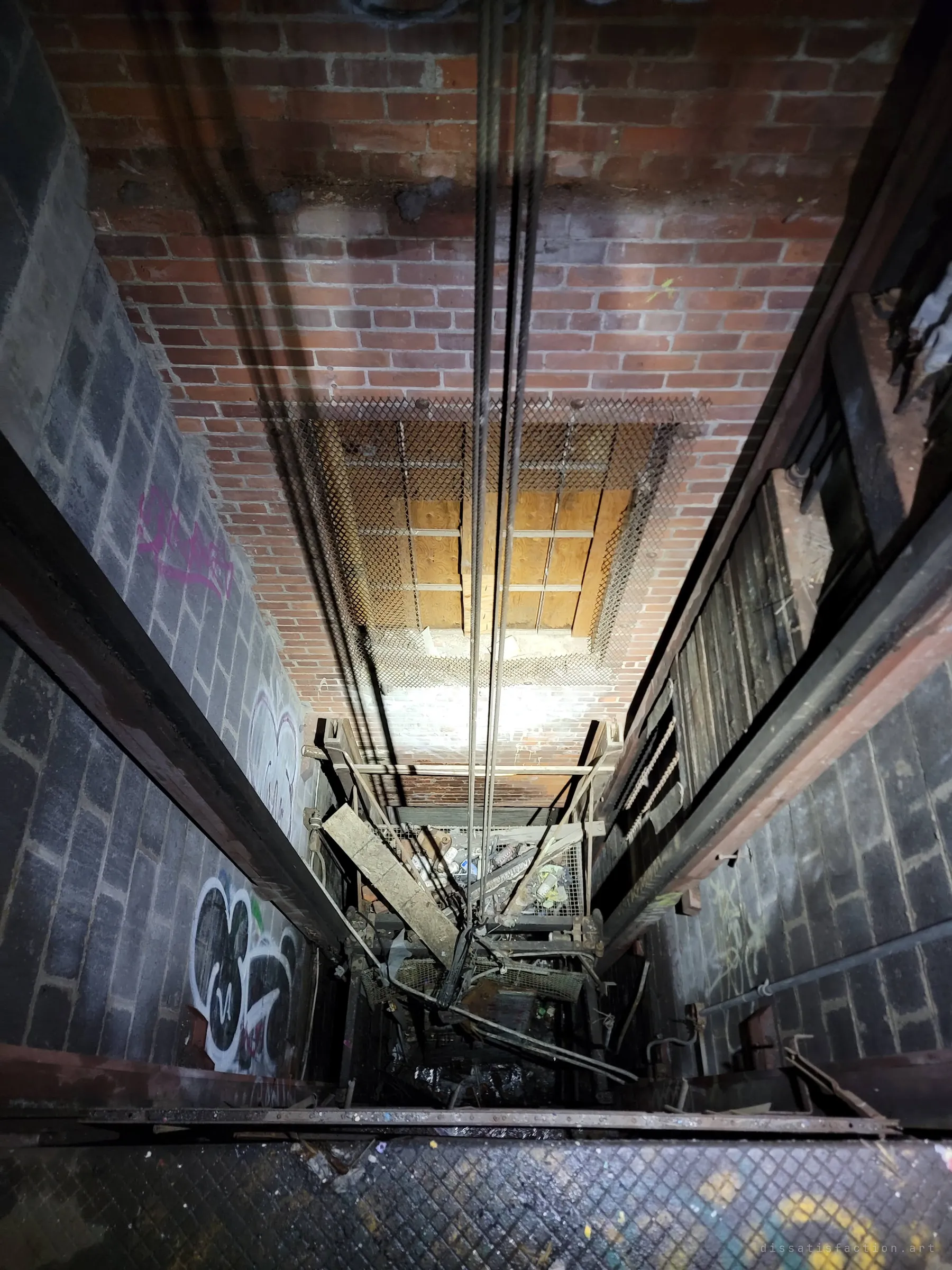 Looking straight down an old freight elevator shaft, cables run past brick and block walls to a wrecked cage heaped with debris at the bottom. The vertigo of the drop is the point of the shot.