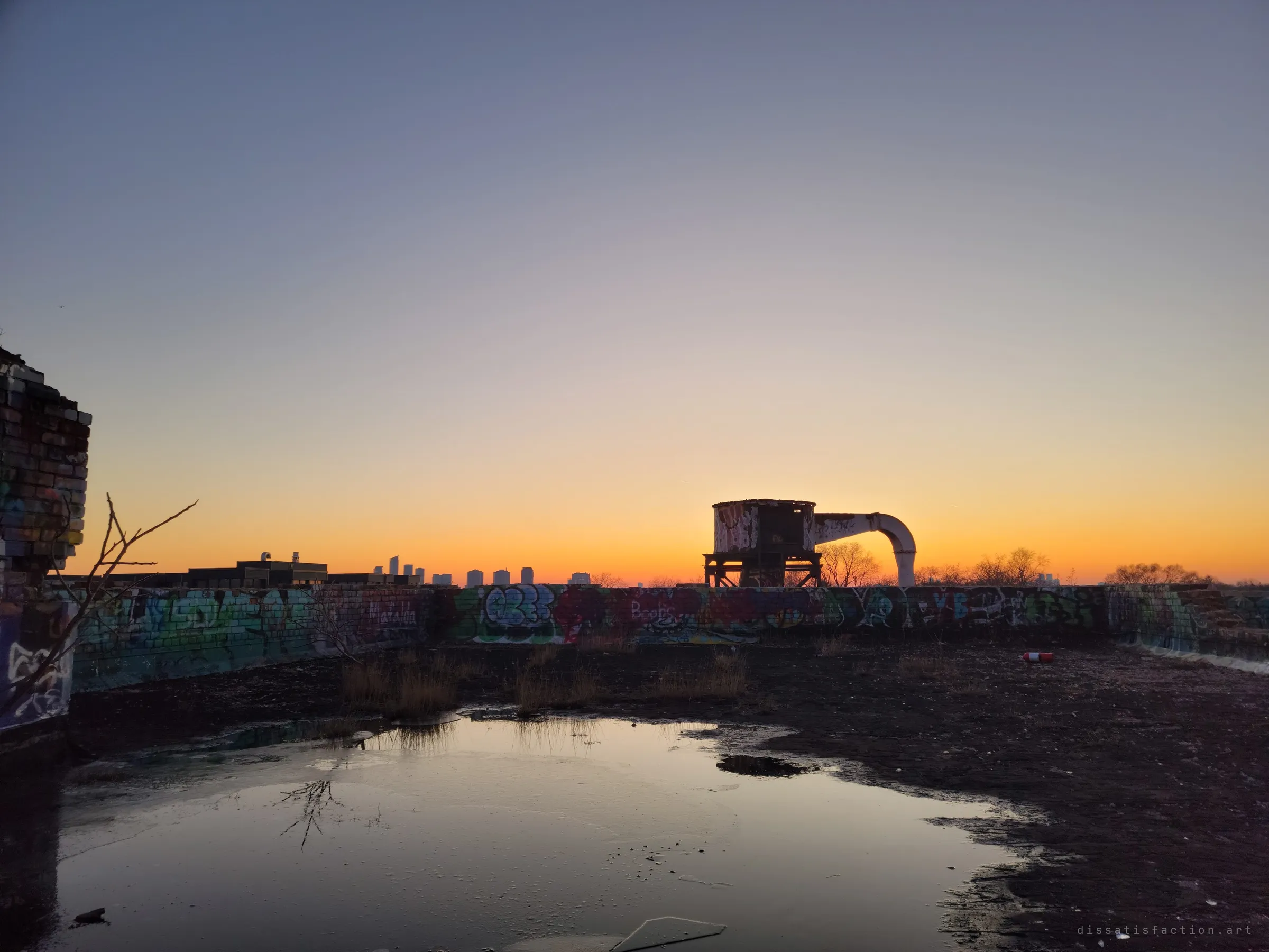 A flooded, graffiti-walled rooftop reflects an orange sunset while an old hopper tank with a hooked chute stands in silhouette. Distant towers sit low on the horizon.