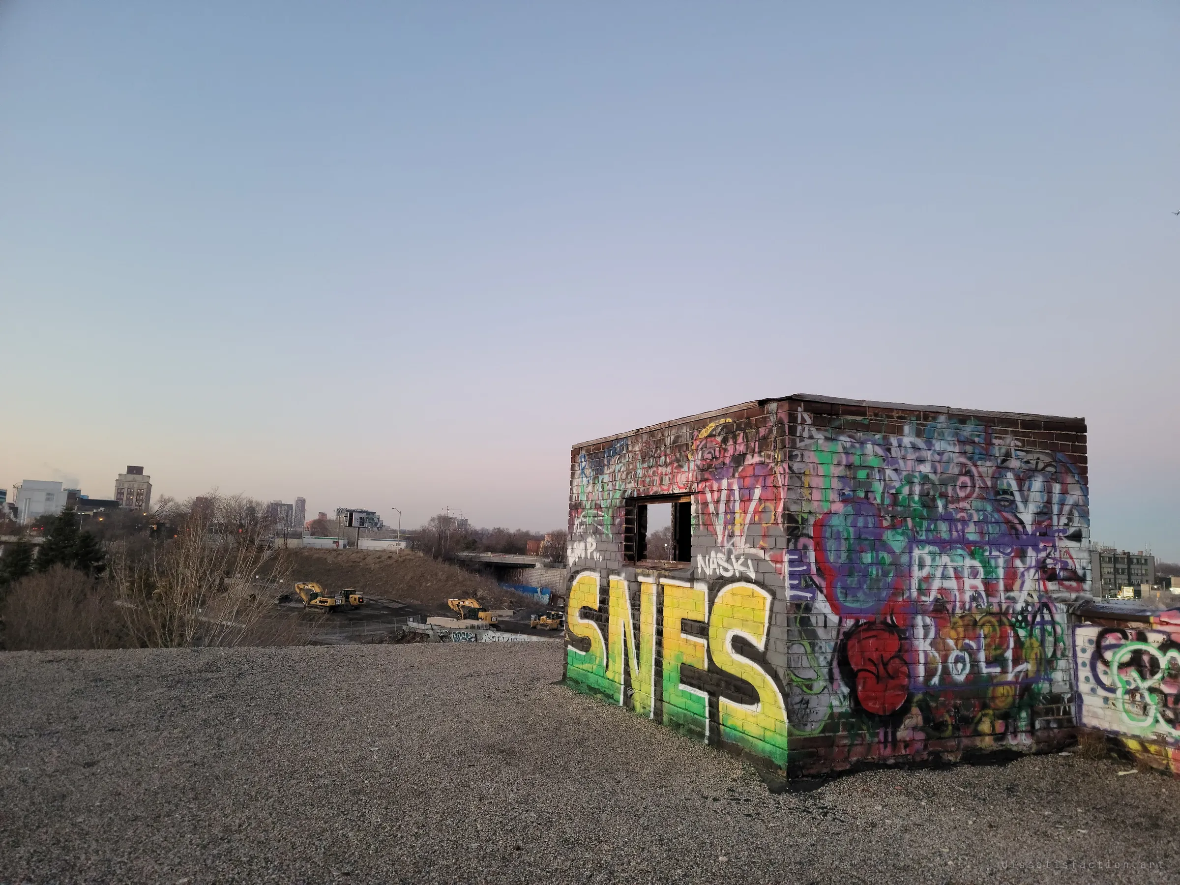 A small brick rooftop hut covered in bright graffiti, with a big yellow-and-green piece on its side, sits on a gravel roof under a pale evening sky. Beyond the edge, excavators work a construction lot.