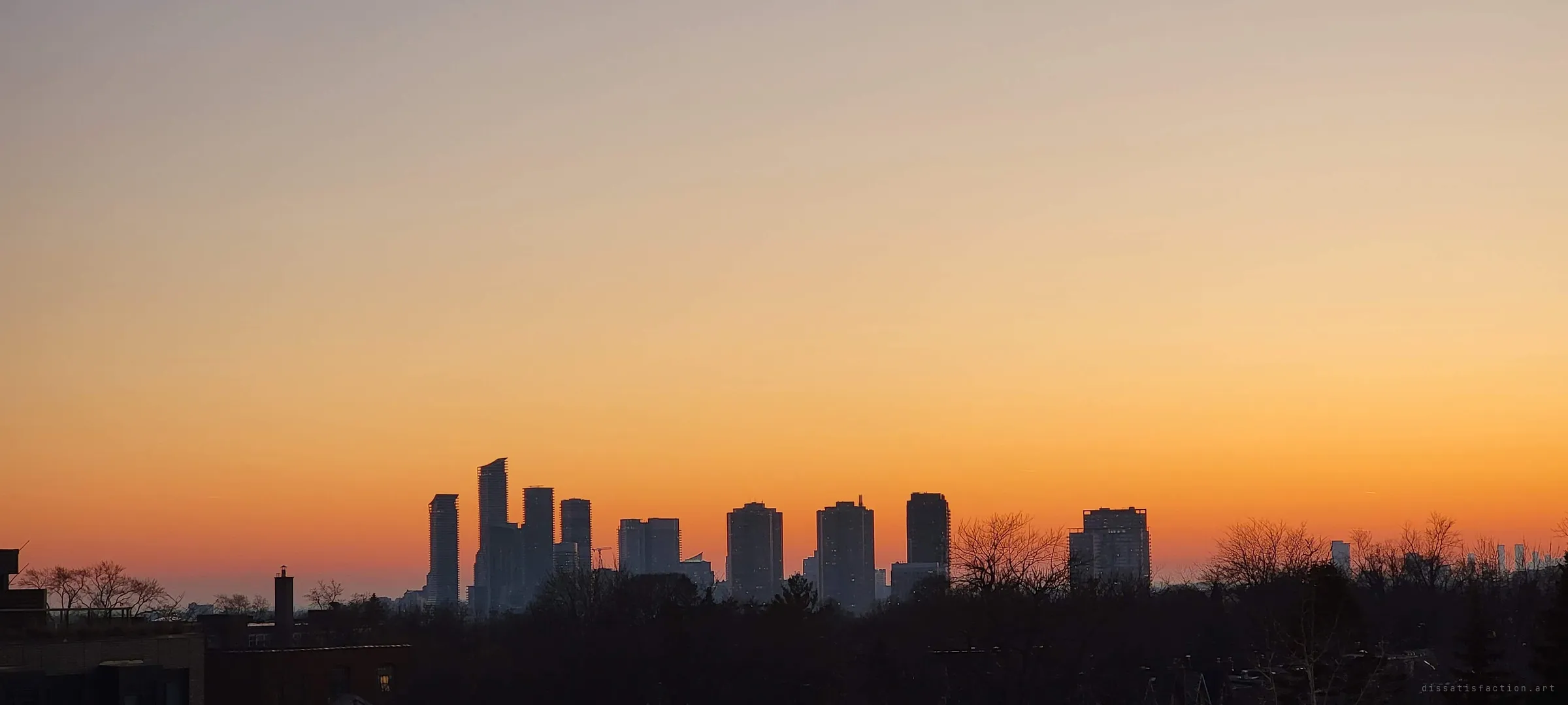 A cluster of high-rise towers stands in silhouette against an orange-to-gold sunset over a dark band of treetops. The view from the roof at golden hour.