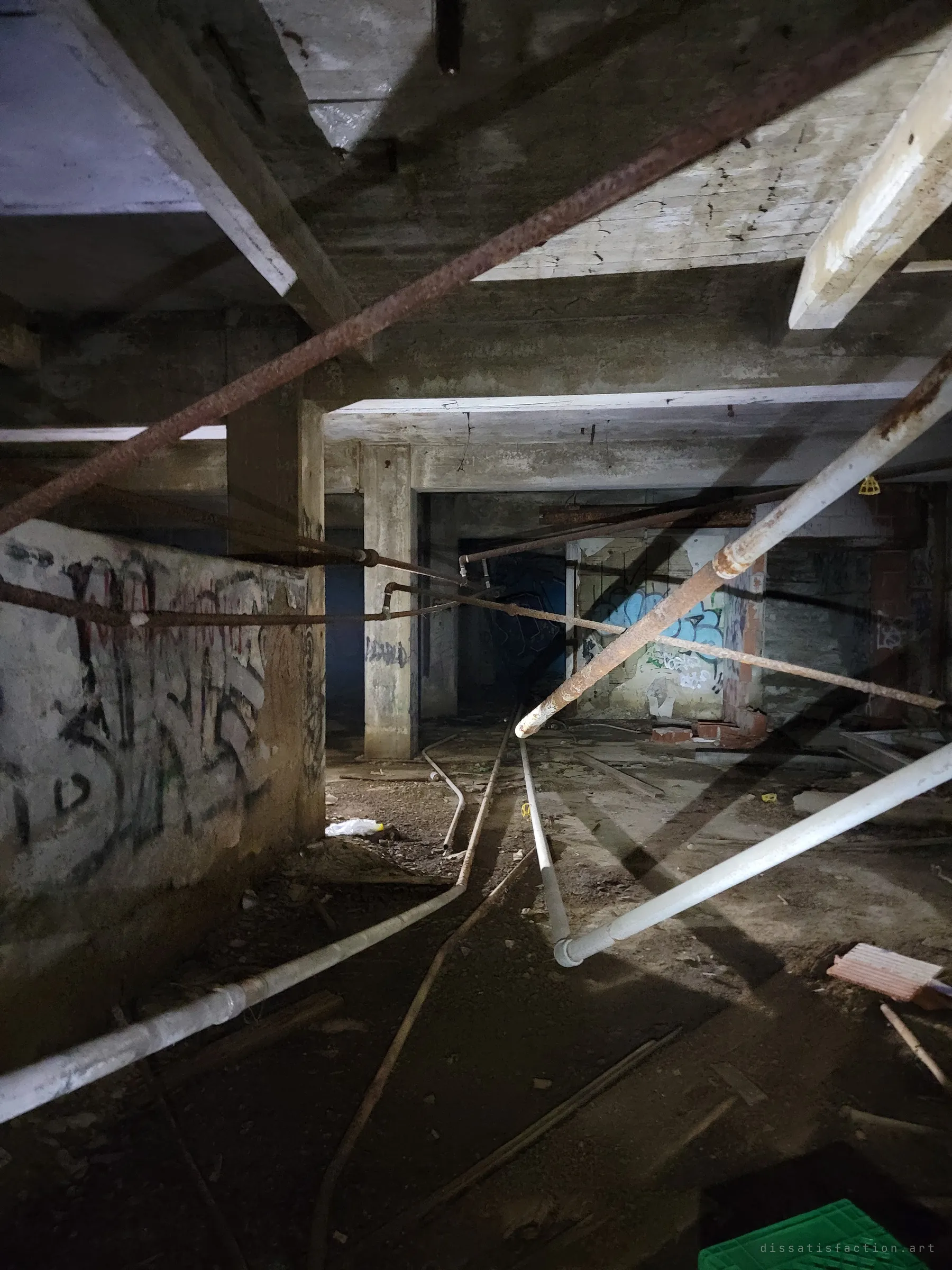 A low concrete basement crossed by sagging rusted and white pipes, with columns and graffiti walls fading into the dark. The flashlight picks out the tangle of old services.