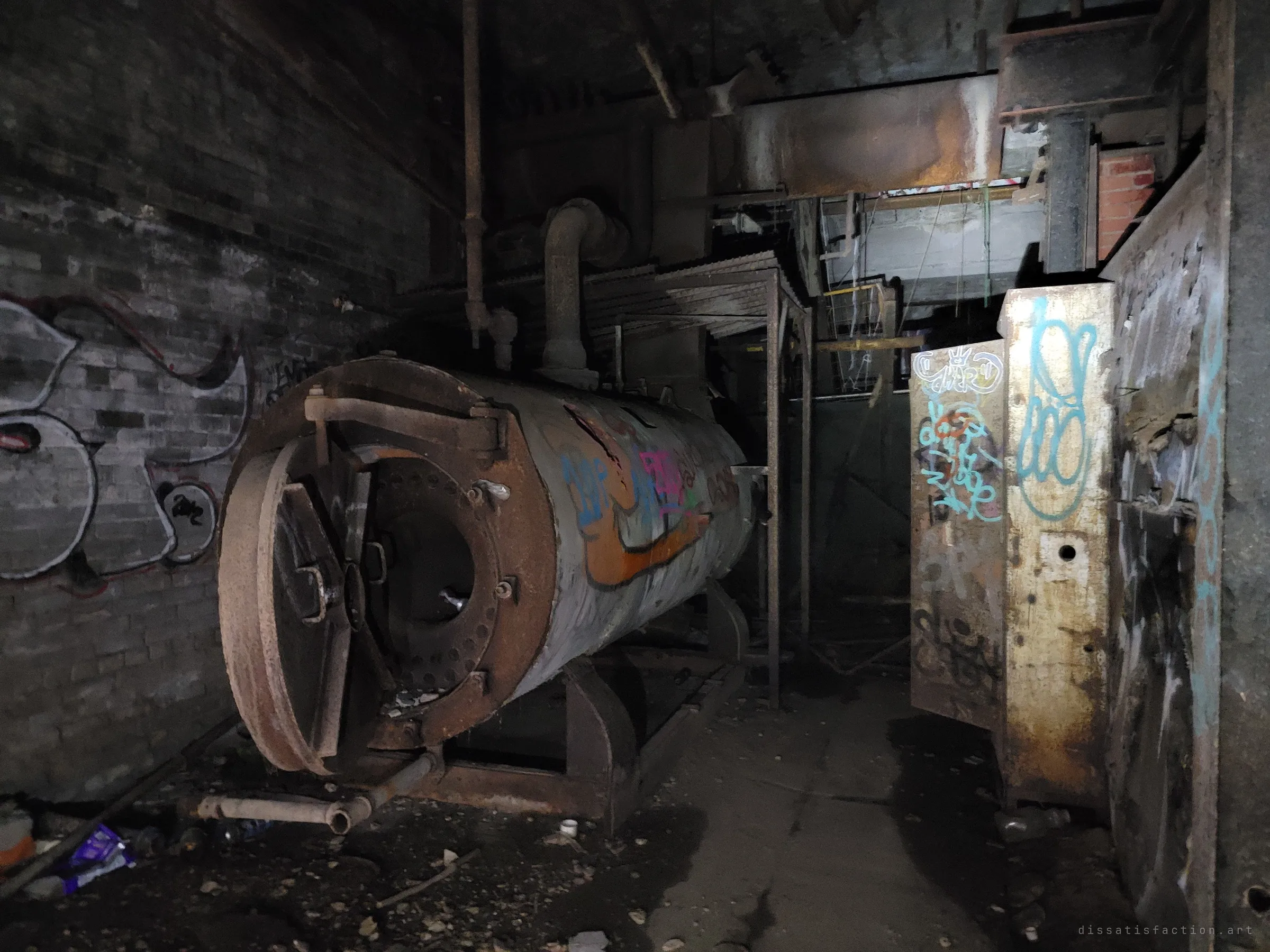 An old horizontal boiler with its rusted front hatch swung open sits in a dark, graffiti-marked boiler room beside tagged steel panels. The heart of the plant, long gone cold.