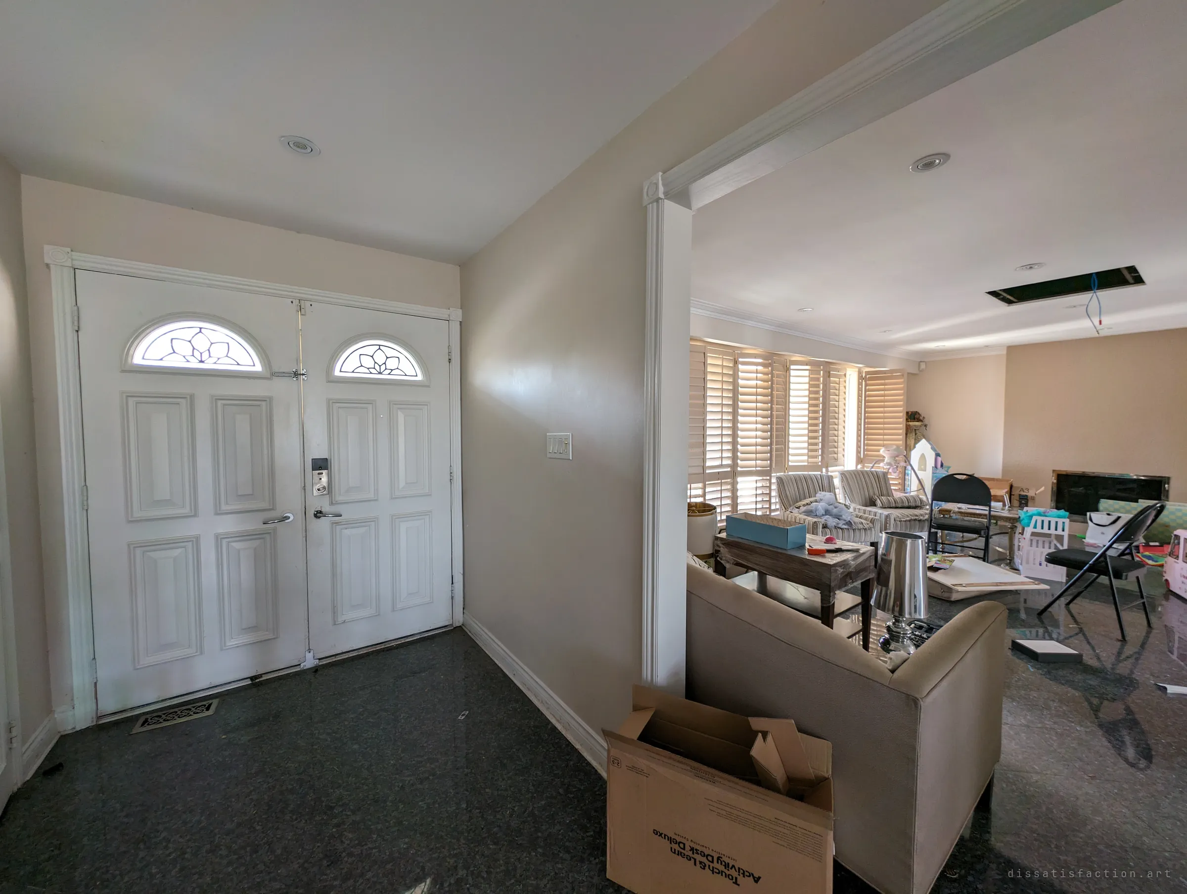 The double front doors of an abandoned house open onto a dim foyer, with a living room of toys, boxes and furniture left behind through the archway. The half-lived-in mess hints at a family that left in a hurry.
