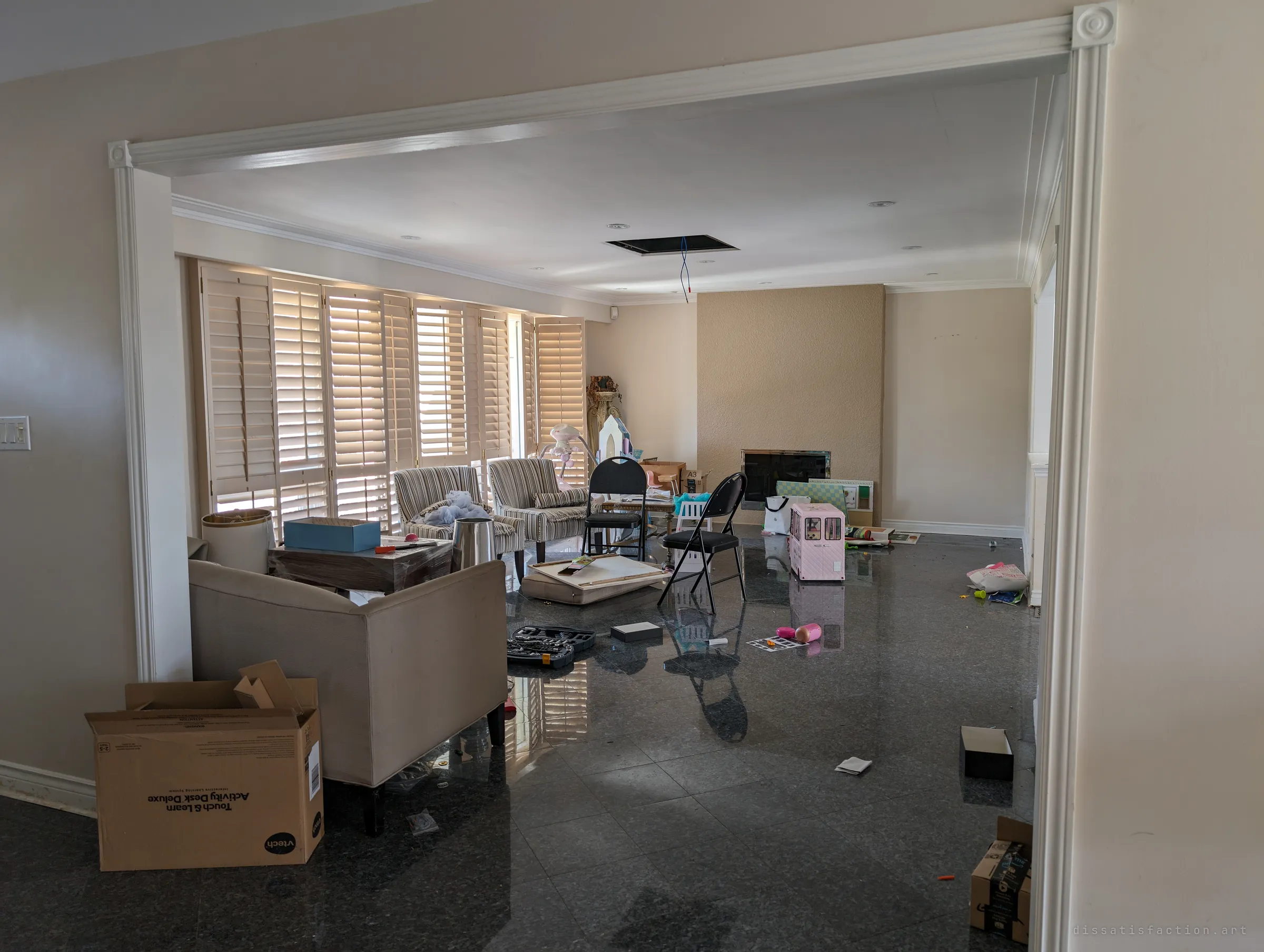 A shuttered living room seen through a white archway, its polished stone floor strewn with toys, a toolkit, folding chairs and a children's playhouse. An open ceiling hatch with dangling wires shows the house has already been picked at.