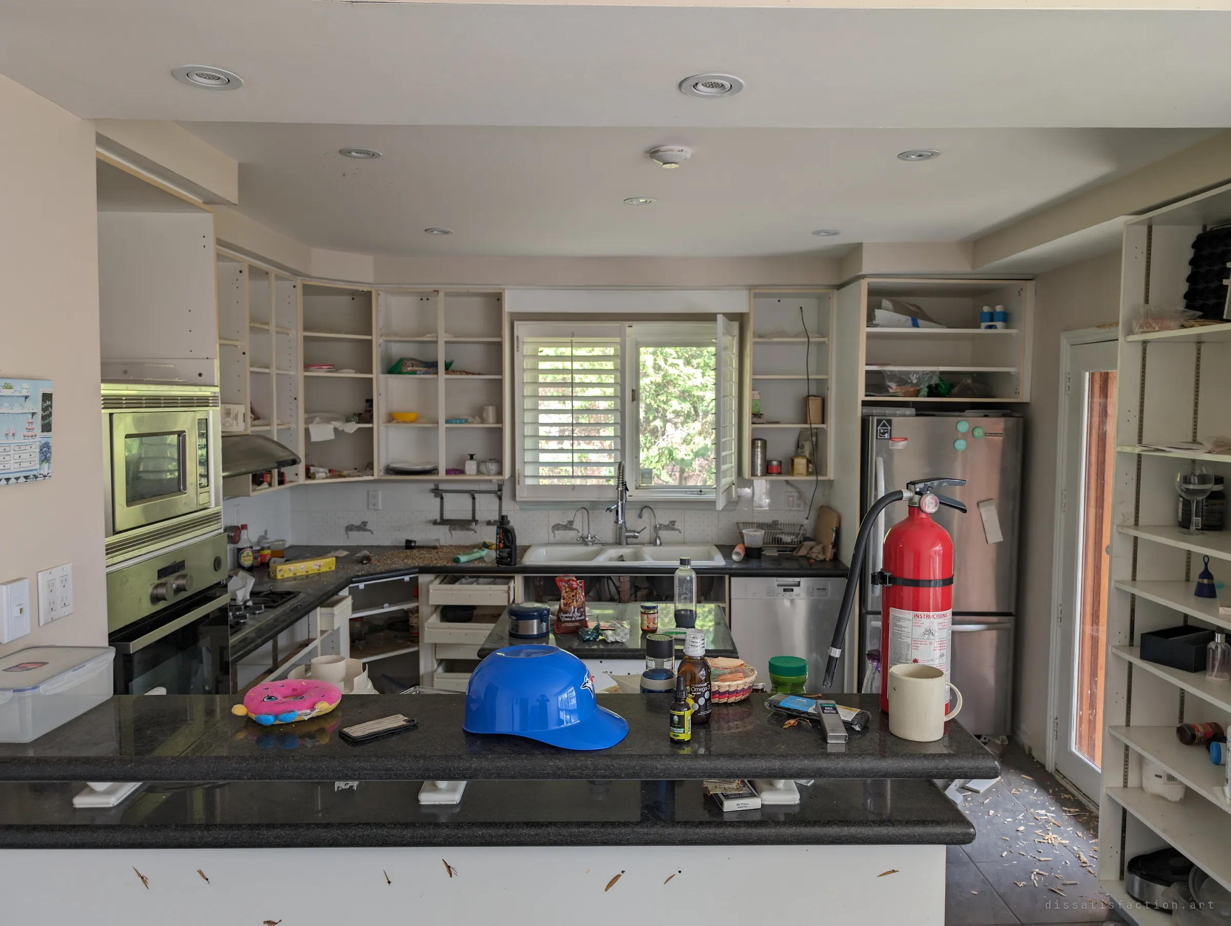 A kitchen with its cabinet doors torn off, the counter still holding a toy helmet, a plush doughnut, supplements and a mug beside a fire extinguisher. Everyday objects sit where they were dropped, as if the owners stepped out and never returned.
