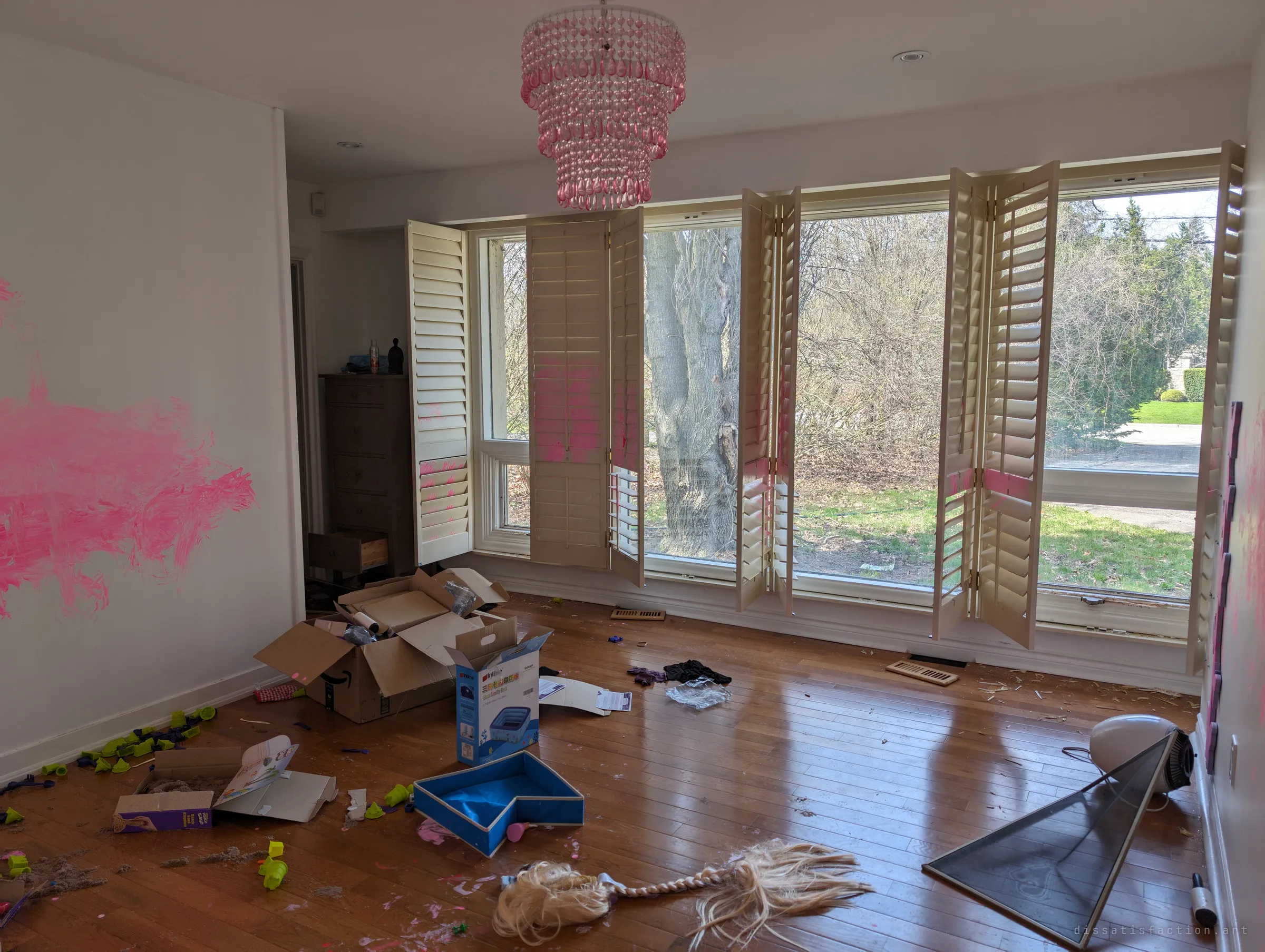 A child's bedroom with pink paint smeared across the wall and shutters, a pink bead chandelier, a discarded blonde wig and toys on the hardwood floor. Tall windows let in bare spring light from the yard.