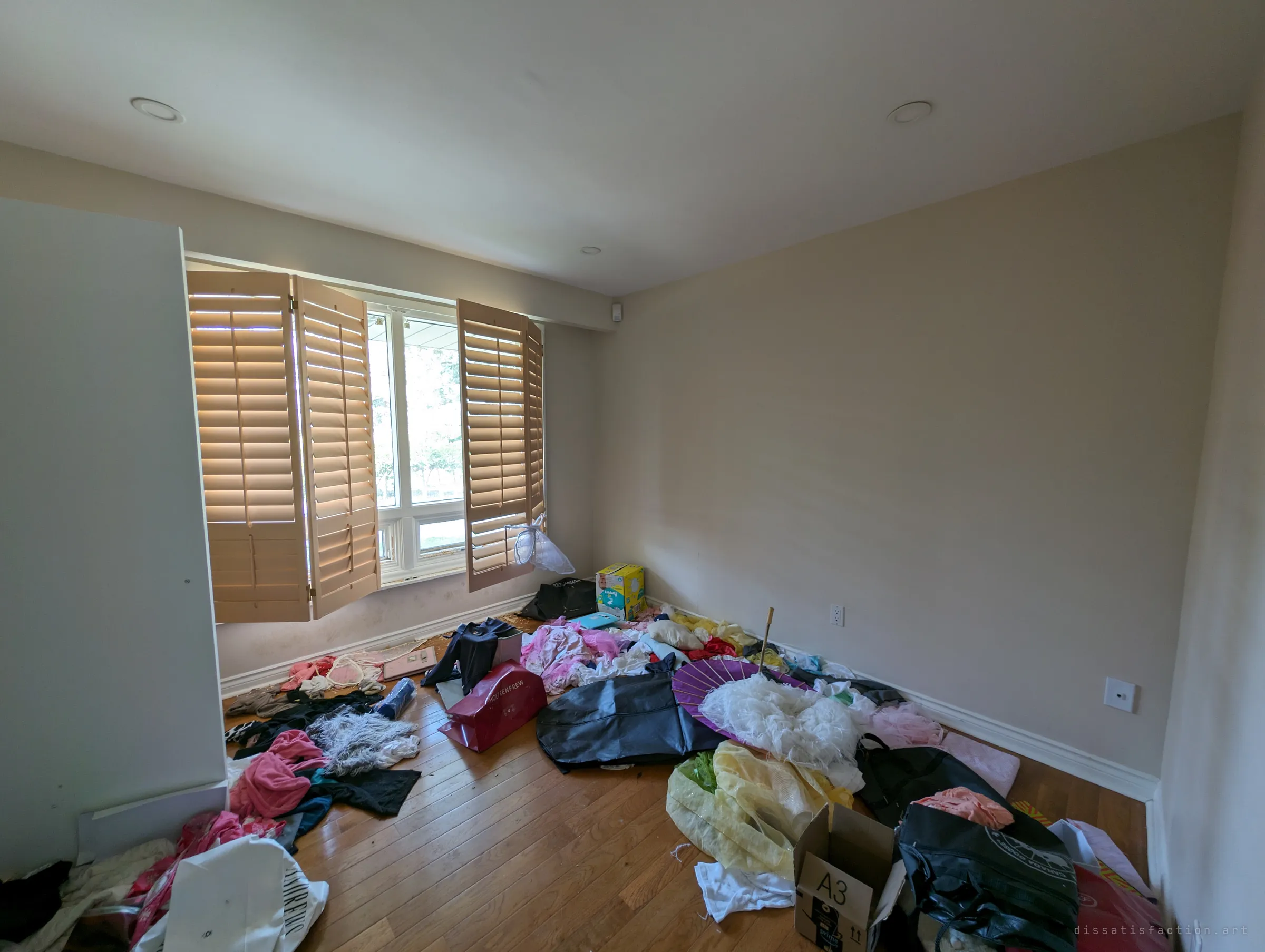 A bedroom with wooden shutters thrown open, the hardwood floor buried in clothes, dresses, bags and boxes. Someone's wardrobe emptied across the floor, as if the room was left mid-packing.