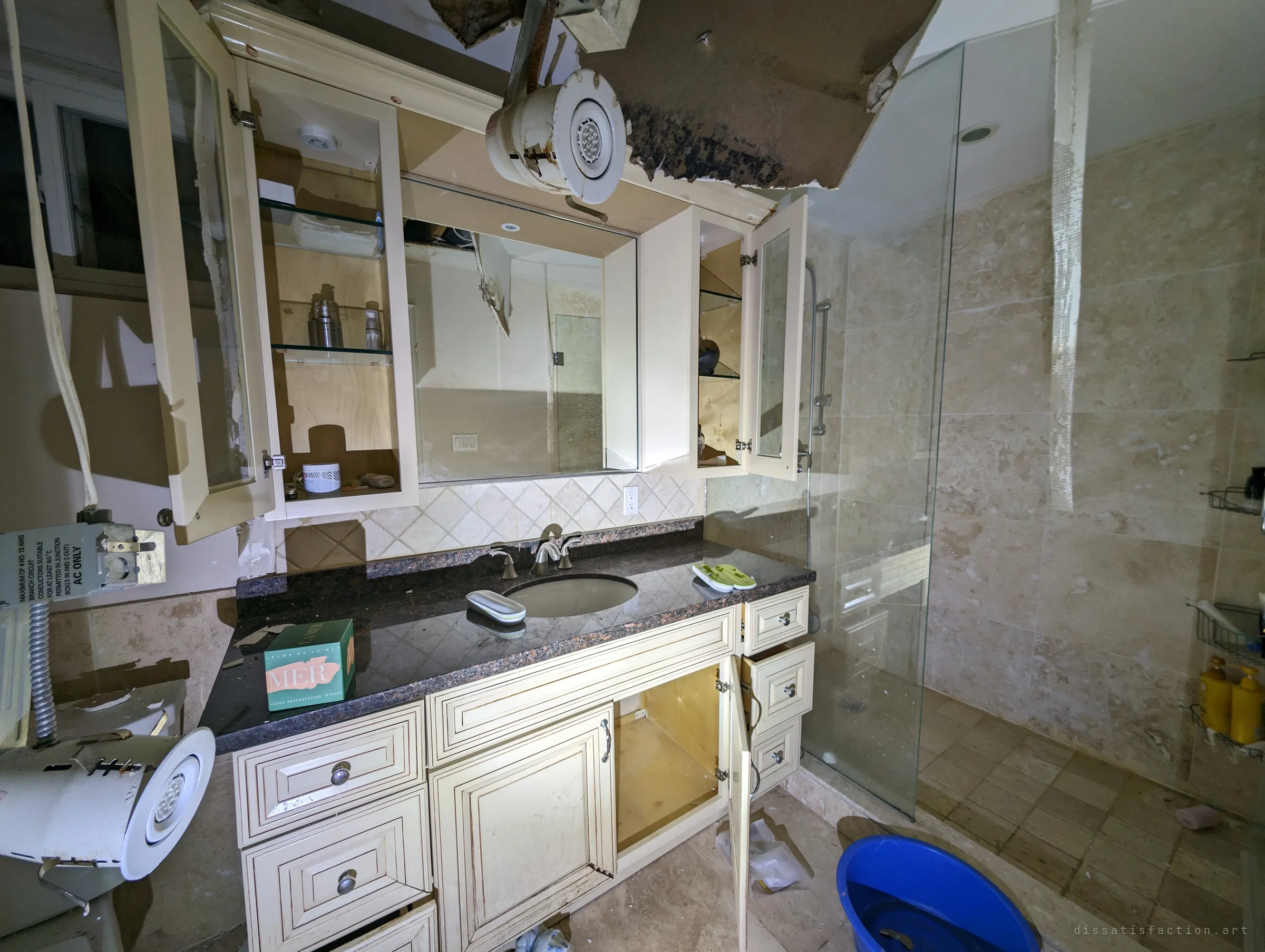 A bathroom vanity with every cabinet door hanging open under a collapsed, mould-stained ceiling, a light fixture dangling from the hole. Toiletries still sit on the shelves and a blue bucket waits by the glass shower.
