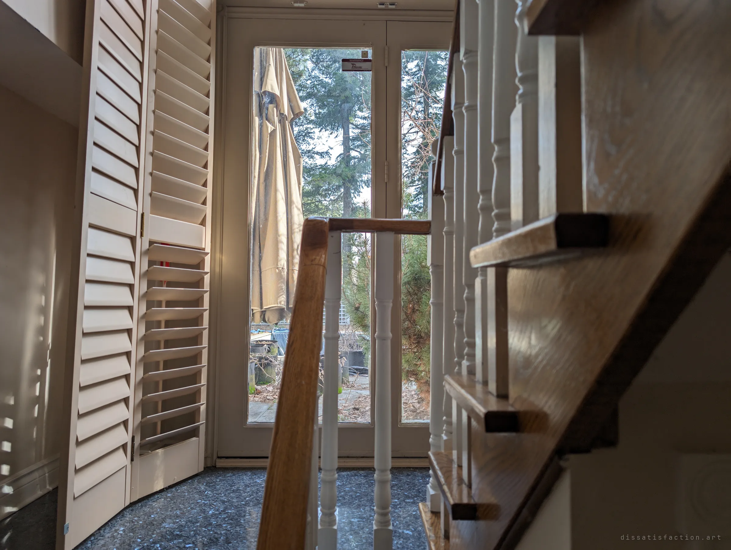 Looking past a wooden banister and folding shutters to a glass back door, a folded patio umbrella and tall evergreens outside. Soft daylight through the slats makes the empty hall feel quietly lived-in.