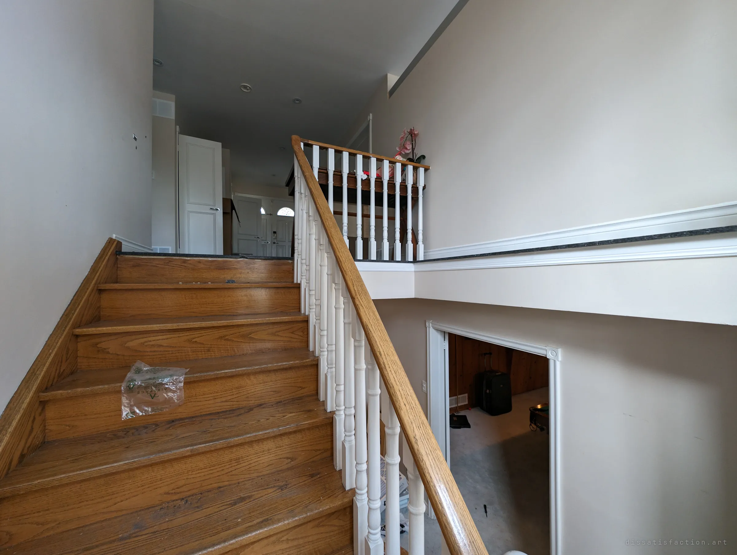 An oak staircase with white spindles rising to the front entry, a lone plastic bag on one step and a suitcase visible in the panelled room below. The house is split between clean upper floors and a cluttered basement.