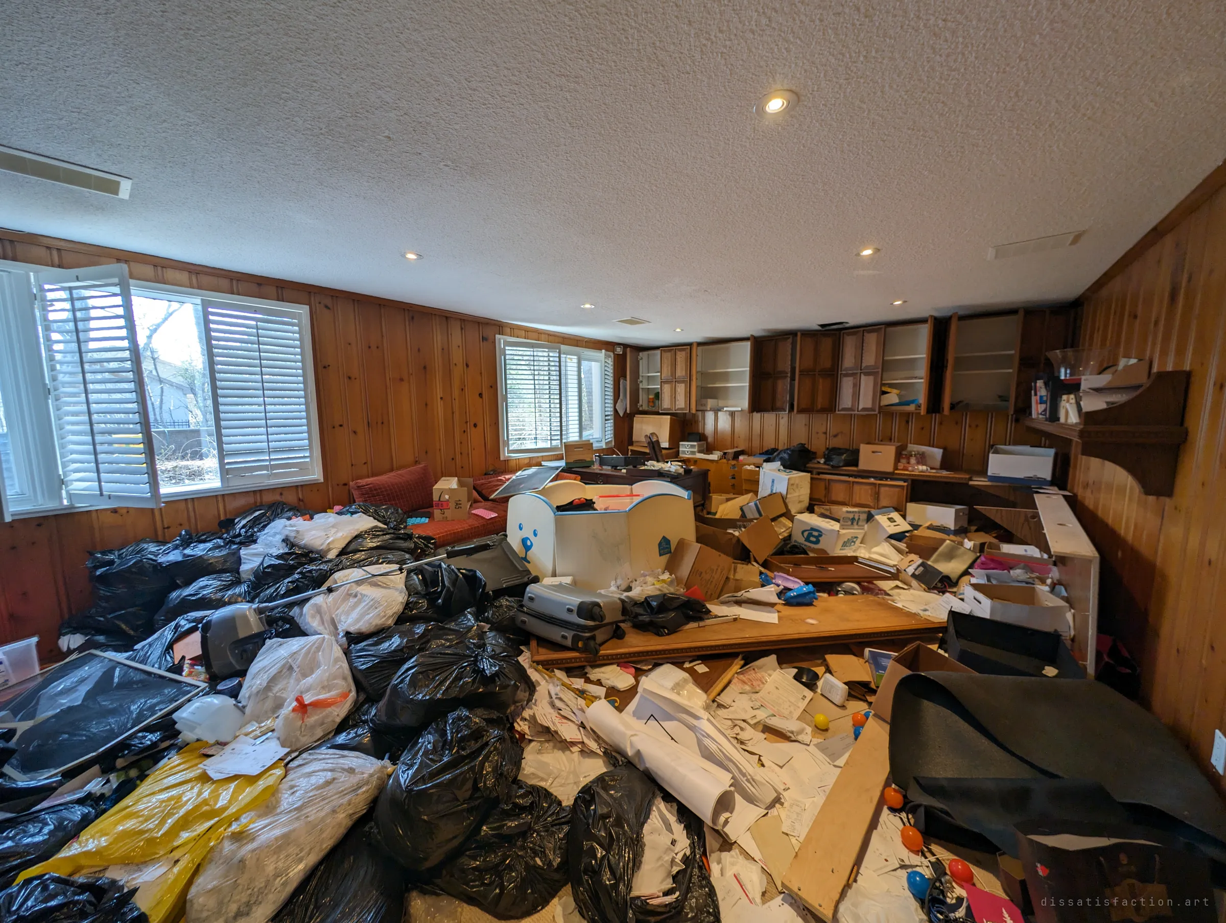 A wood-panelled basement room drowned in black garbage bags, suitcases, boxes and drifts of paperwork, with every cabinet on the far wall opened. It looks like a life was bagged up and abandoned halfway through.