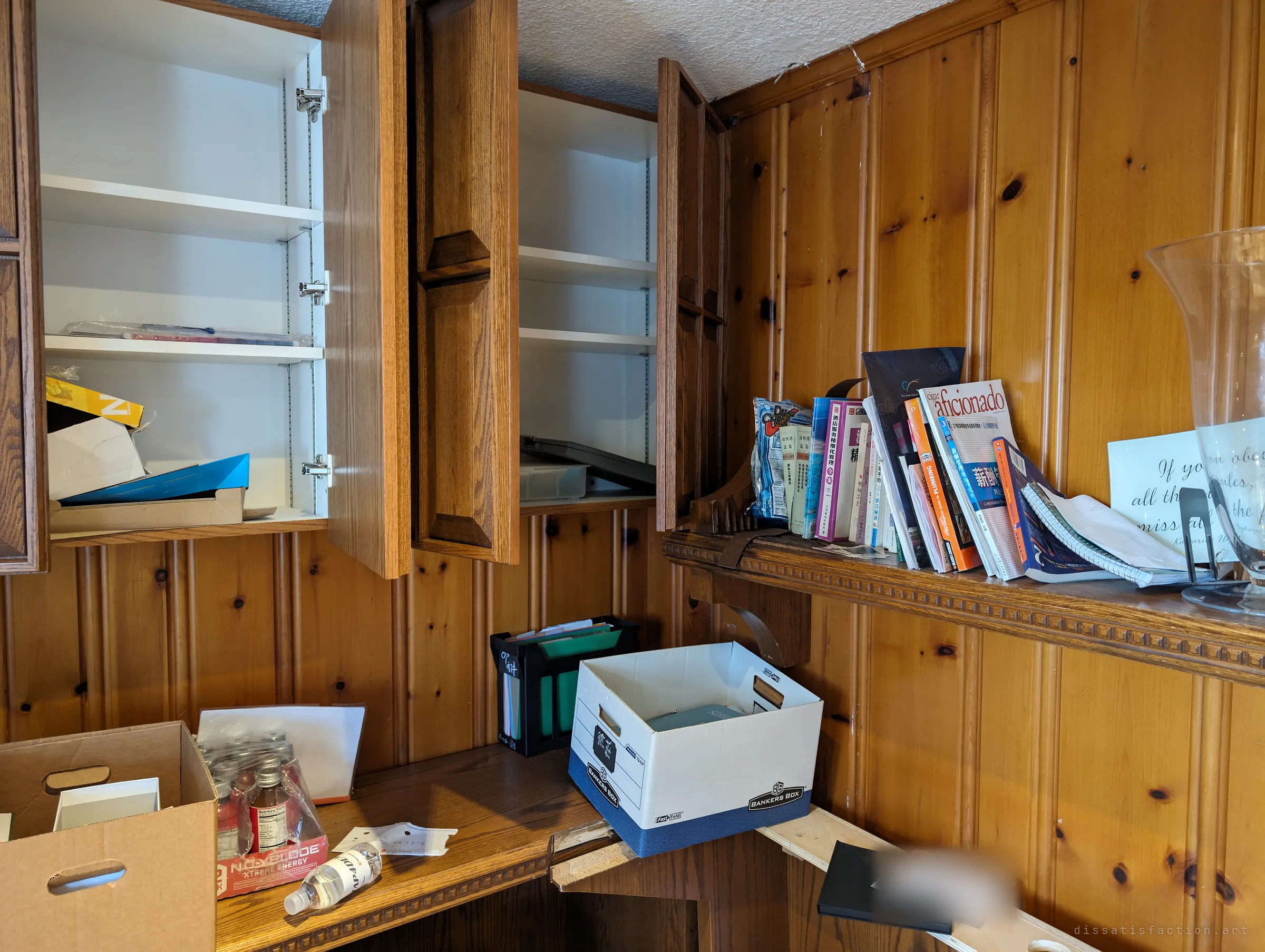 Open cabinets above a panelled desk, a shelf of Chinese and English books, a banker's box and a crate of energy drinks. Household papers still lie on the desk as if someone stepped away mid-task.