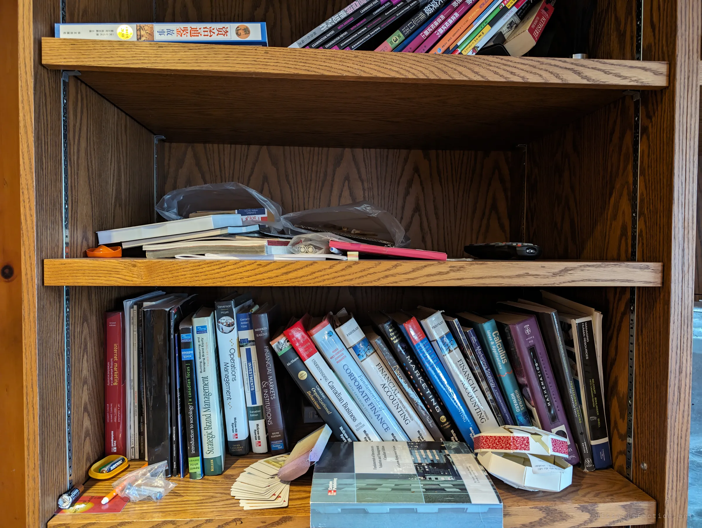 A wooden bookcase of business, finance and accounting textbooks, with Chinese-language books and exam papers on the top shelf. The study material of someone's degree, left behind with a TV remote and a deck of cards.