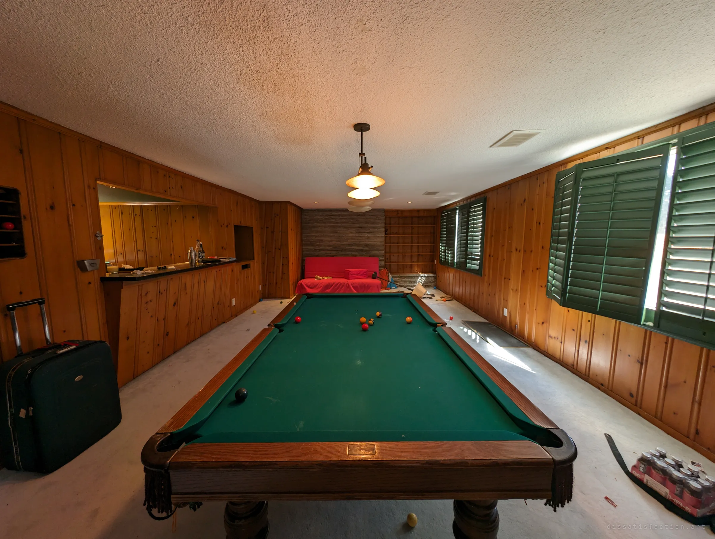 A green-felt pool table centred in a knotty-pine basement, balls still scattered mid-game under a hanging lamp, with a bar on the left and green shutters on the right. A suitcase stands by the table as if packed and forgotten.