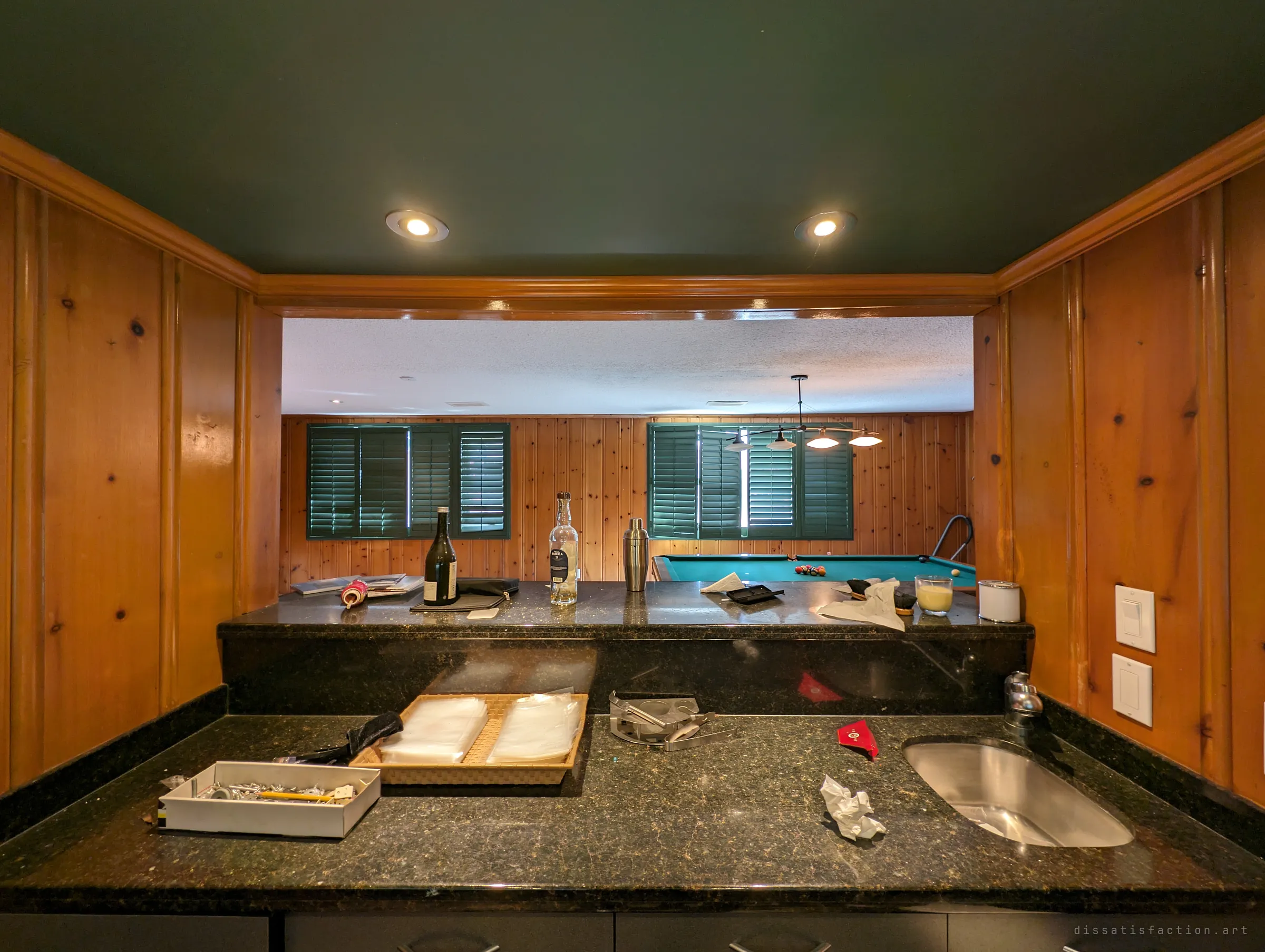 From behind the basement bar, a granite counter with bottles, a cocktail shaker and a sink looks through a pass-through to the pool table and green shutters. The bar is still set as if the last drinks were never cleared.