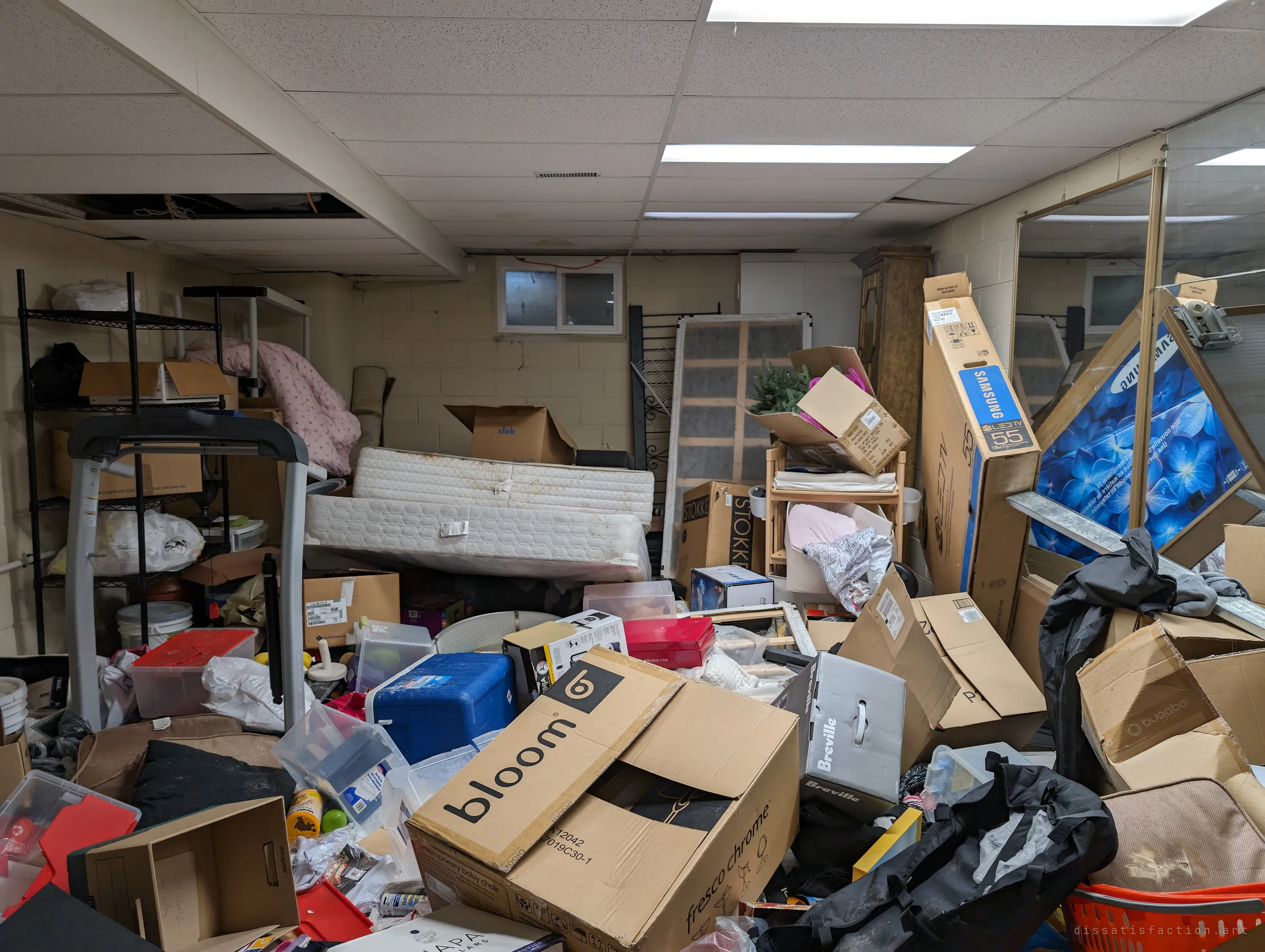 A basement rec room buried to the ceiling in boxed TVs, mattresses, baby gear and a treadmill under drop-ceiling lights. The household's belongings were packed, then simply left.