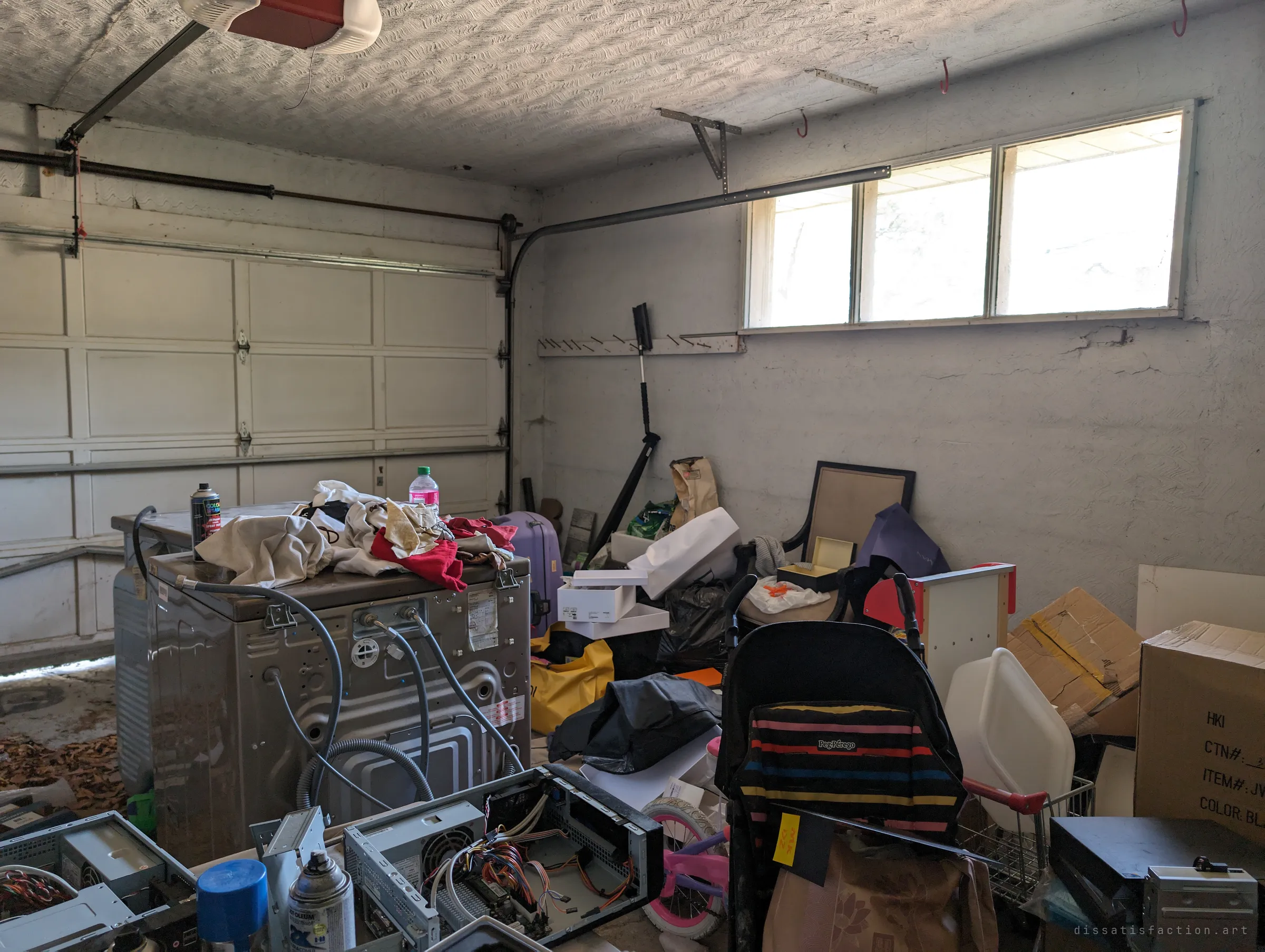 An attached garage with a washing machine pulled out, gutted computer towers, a stroller and a child's bike crowding the closed door. Dead leaves have blown in across the concrete floor.