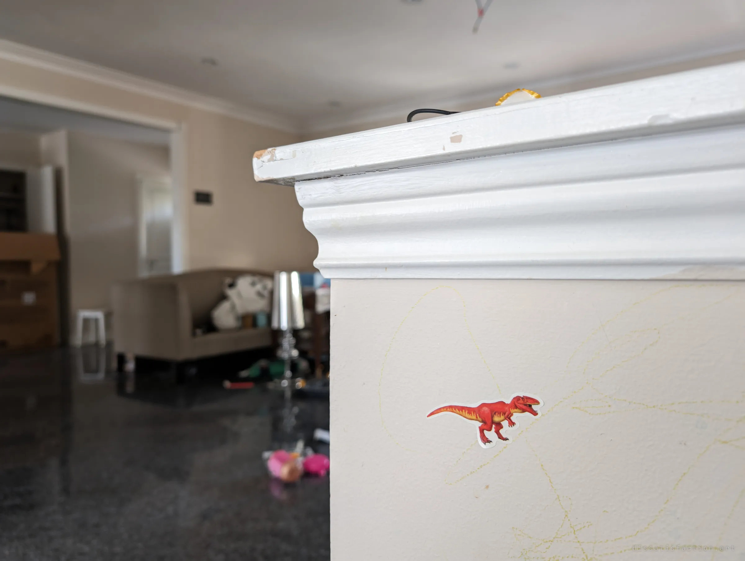 A dinosaur sticker and crayon scribbles on a white pillar, the living room with scattered toys blurred behind it. Small traces of a child left in the empty house.