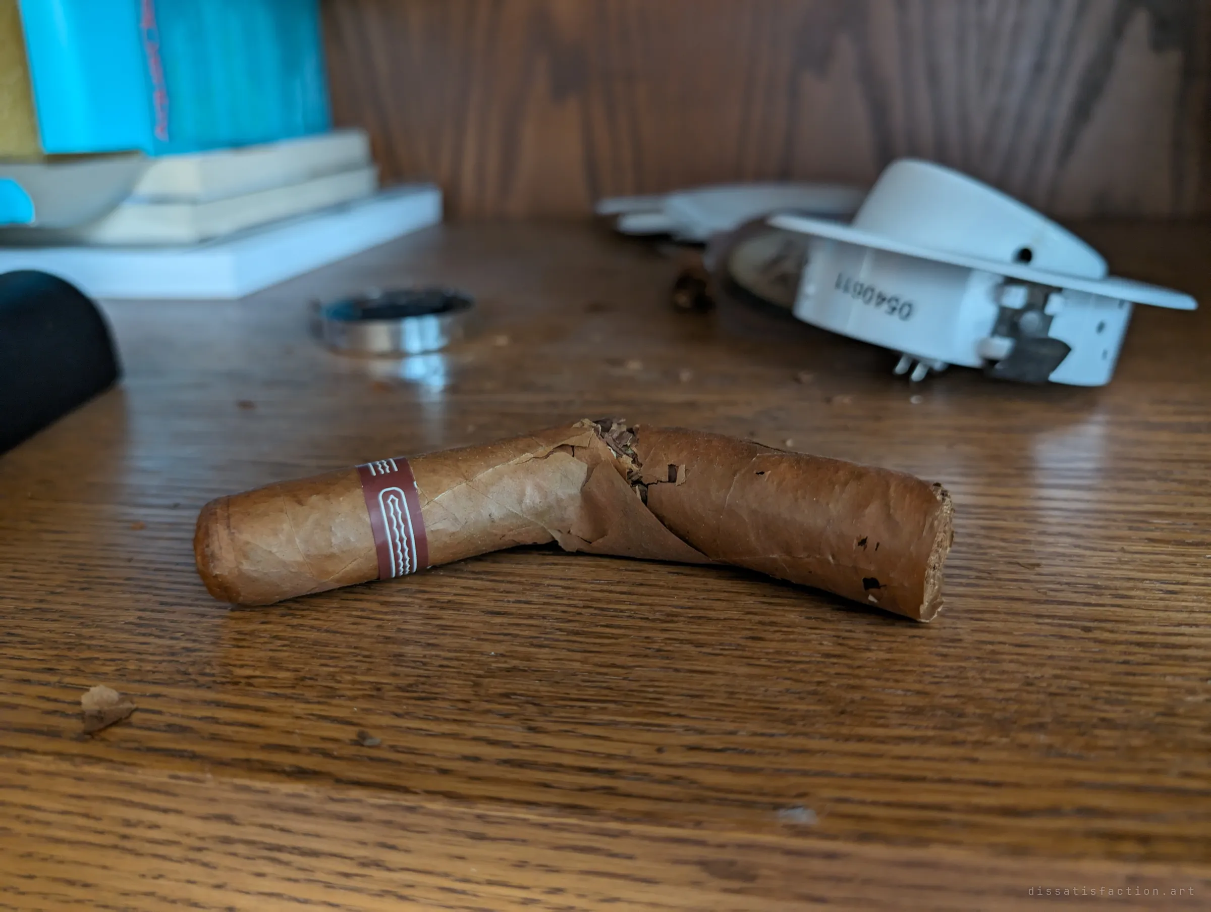 A broken, half-smoked cigar lying on a wooden desk beside a small ashtray and loose tobacco flakes. A personal habit frozen mid-use.