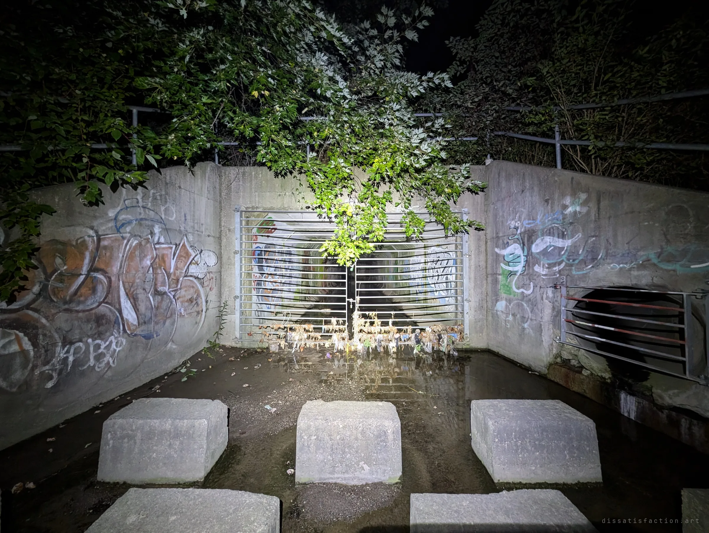 A barred storm drain outfall at night, its concrete headwall layered with graffiti and hung with flood debris, concrete blocks standing in the shallow water in front. The gate is the threshold between the creek and the drain.