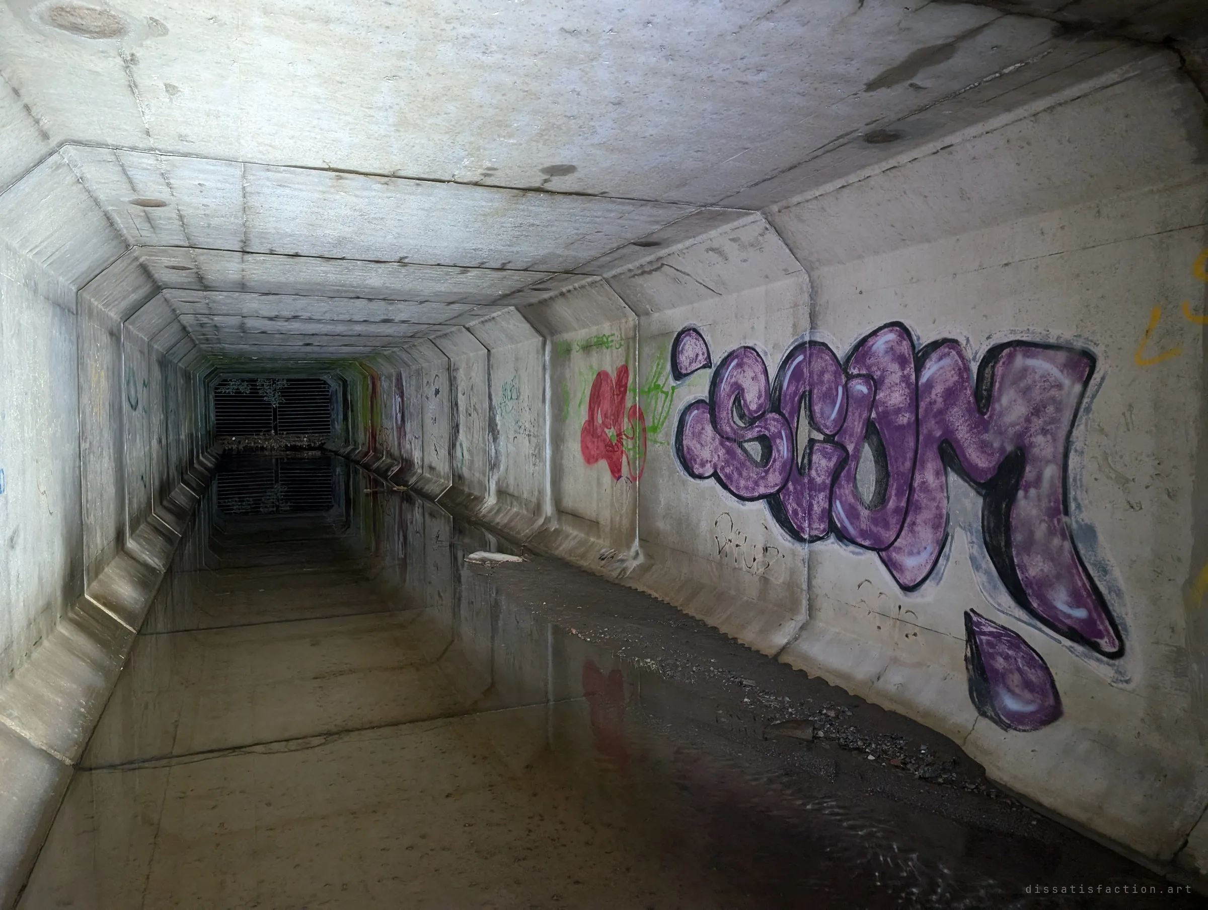 A long precast box culvert running back toward the distant grate, with a large purple graffiti piece on the right wall and still water mirroring the chamfered segments. The repeating joints show how far the drain runs.