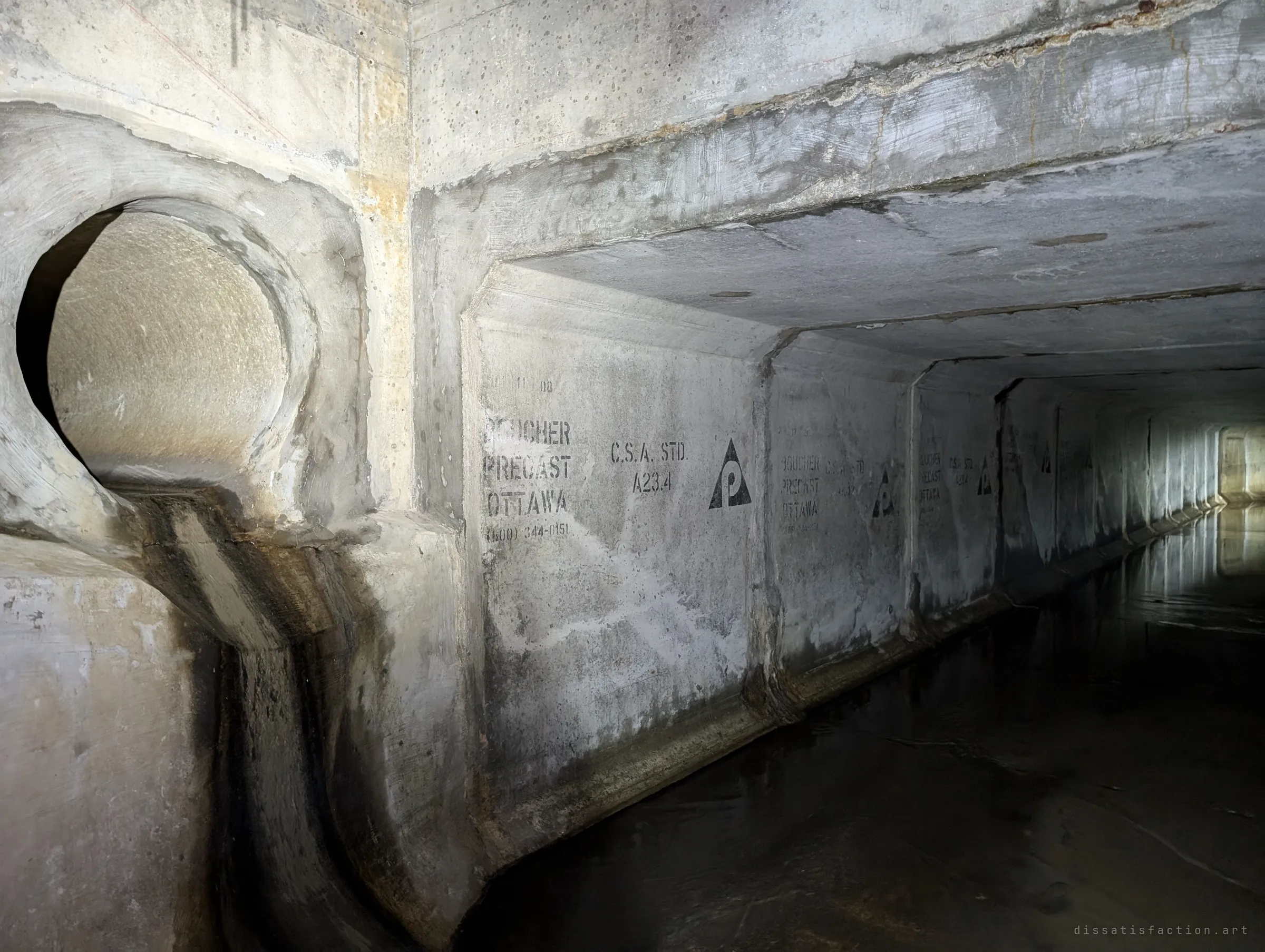 A round pipe joins the box culvert from the side, its outflow having stained a dark channel down the concrete, while stencilled manufacturing marks repeat along the segments into the distance. The markings date and certify each precast section.